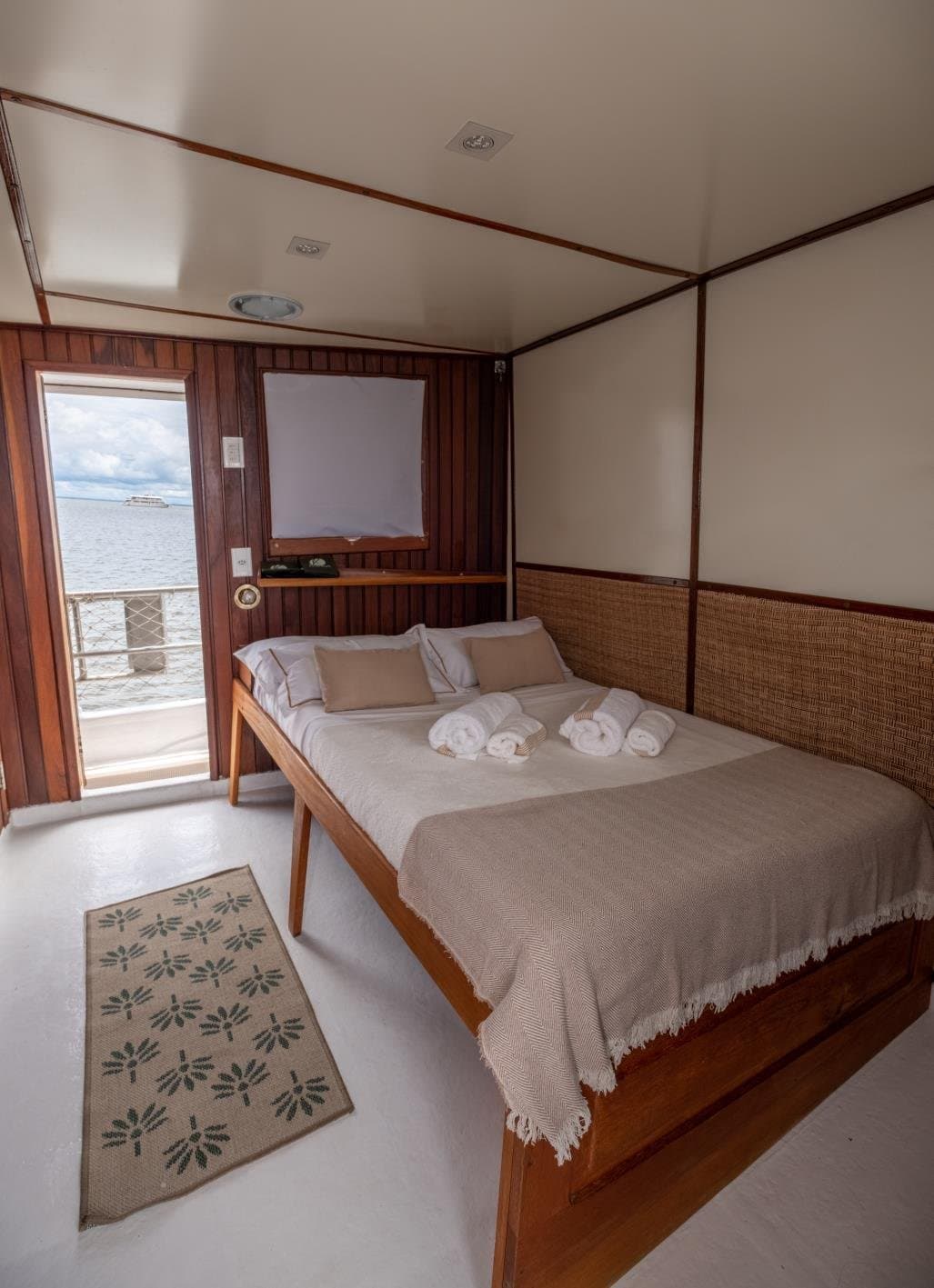Schlafen, während der Amazonas vorbeizieht, das klingt besser als jedes Hotel. Ein ordentlich gemachtes Doppelbett mit gerollten weißen Handtüchern in einer Kabine mit Rattankopfteil an Bord einer Amazonas-Kreuzfahrt, durch die offene Tür ist ein ruhiger Flussblick zu sehen