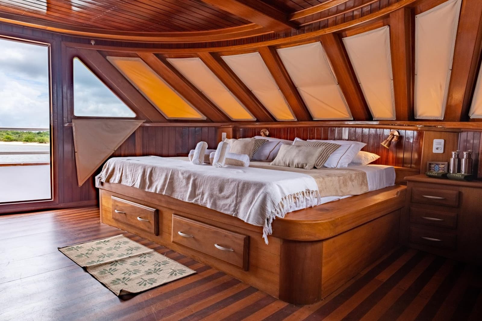 Gemütlicher schlafen als erwartet, mitten im Amazonas. Gepflegte Holzkabine auf einem Amazonas-Kreuzfahrtschiff ab Alter do Chão mit eingebautem Doppelbett, Schubladen-Stauraum, Dachluken aus Segeltuch und einem offenen Seiteneingang mit Blick auf den Fluss