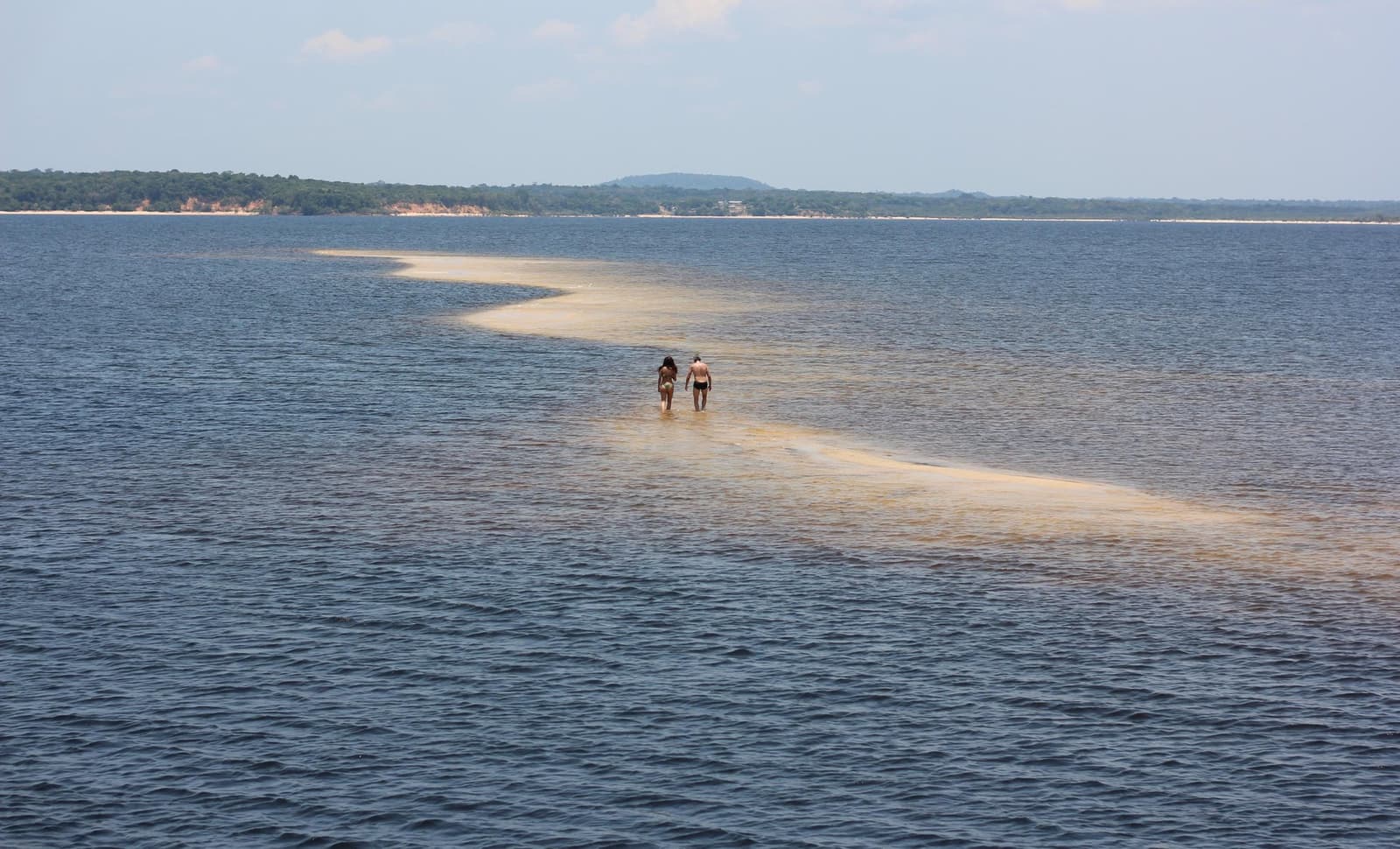 Eine Sandbank, die der Fluss nur gelegentlich freigibt. Zwei Reisende waten entlang einer schmalen Sandbank, die sich über breite blaue Flusswasser erstreckt, auf einer Amazonas-Kreuzfahrt ab Alter do Chão, mit bewaldetem Ufer und sanften Hügeln im Hintergrund