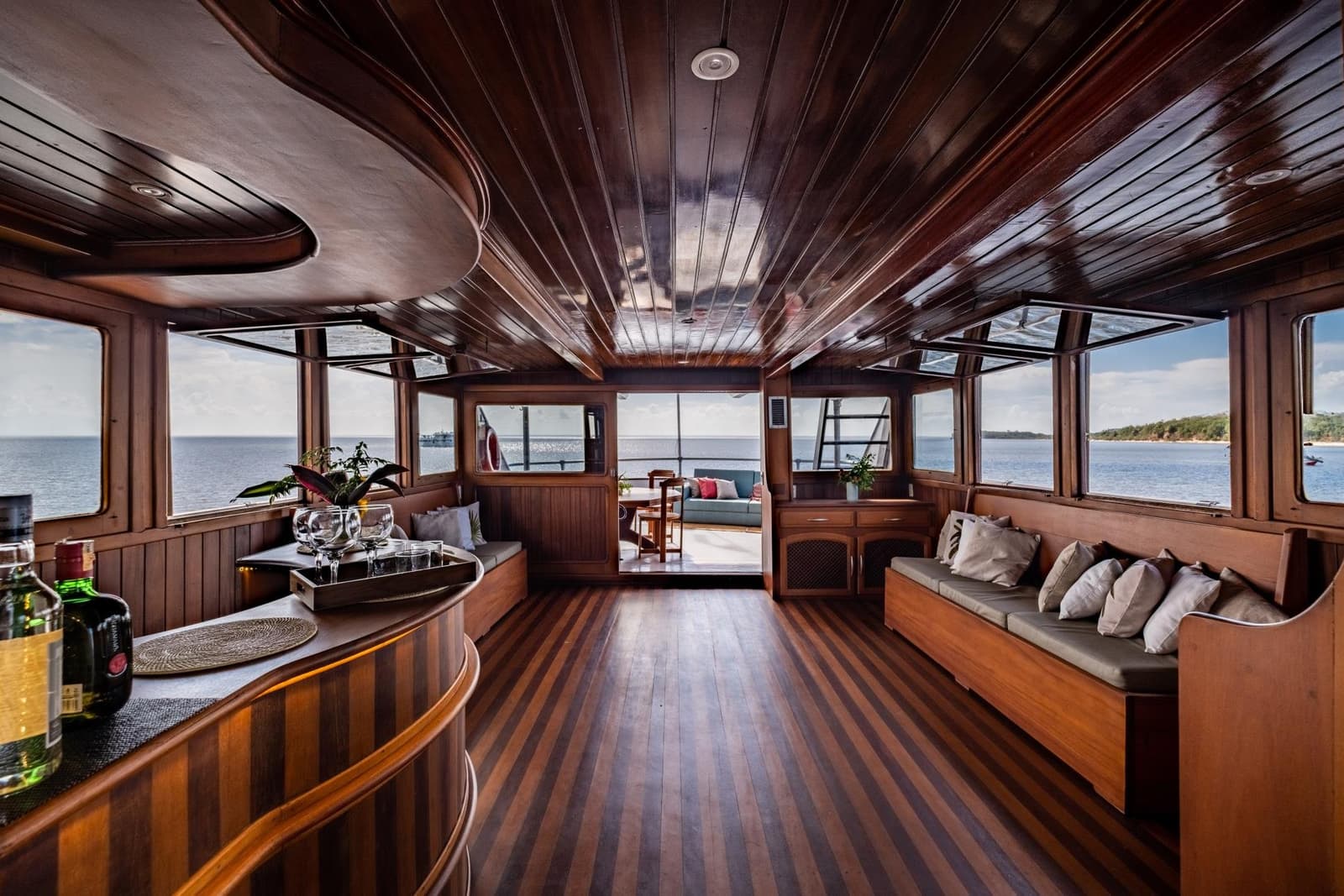 Ein Wohnzimmer auf dem Wasser, nur mit besserem Ausblick. Holzvertäfelter Salon an Bord einer Amazonas-Kreuzfahrt ab Alter do Chão mit eingebauten Polstersitzen, einem Tablett mit Gläsern an der Bar, gestreiftem Teakboden und Panoramablick auf den Fluss durch umlaufende Fenster