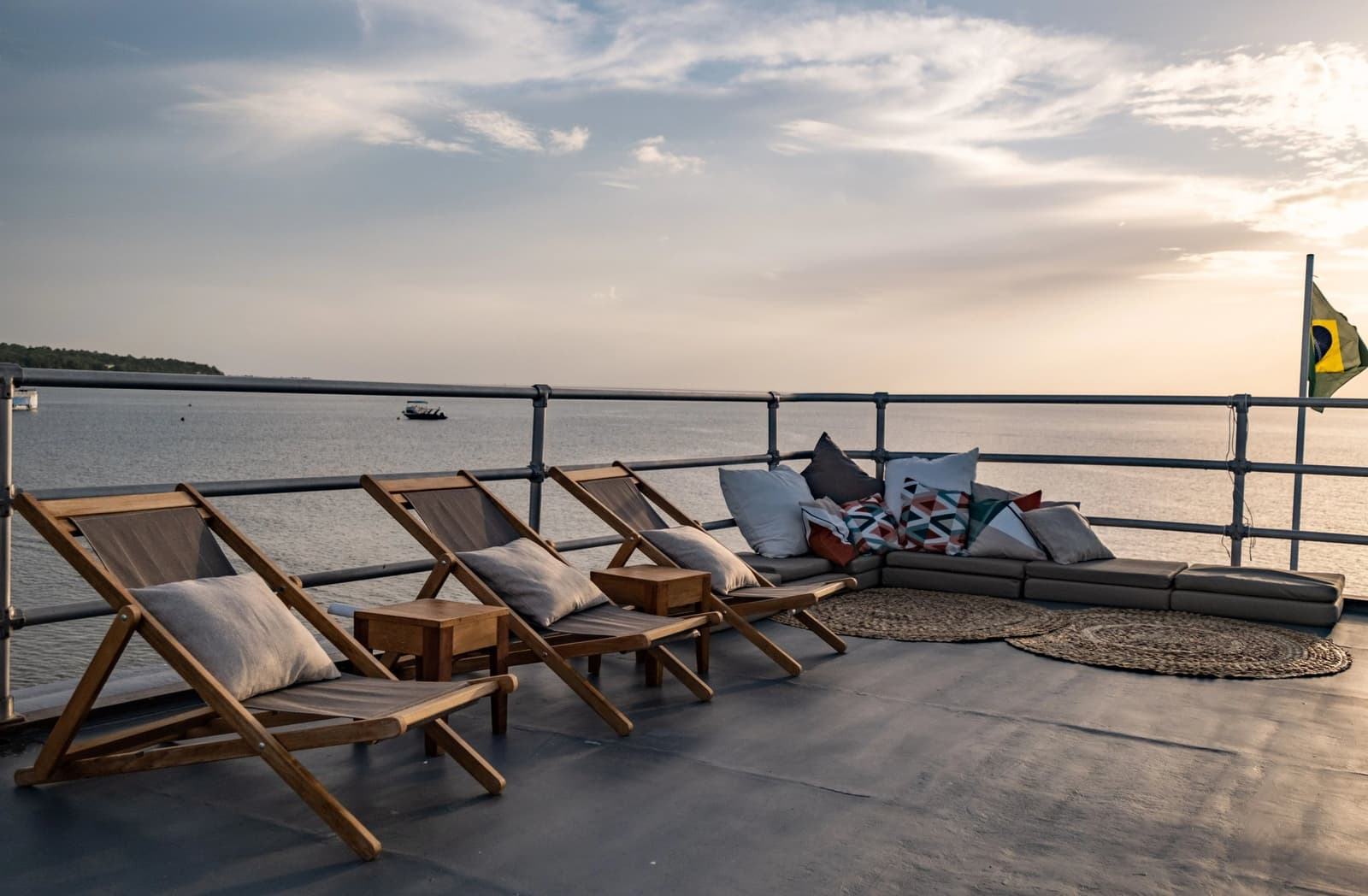 Kein schlechter Platz, um den Amazonas vorbeiziehen zu lassen. Teakholz-Liegestühle und eine gepolsterte Lounge-Ecke auf dem Oberdeck einer Amazonas-Kreuzfahrt ab Alter do Chão, mit einem breiten Fluss unter einem sanften Abendhimmel und der brasilianischen Flagge am Heck