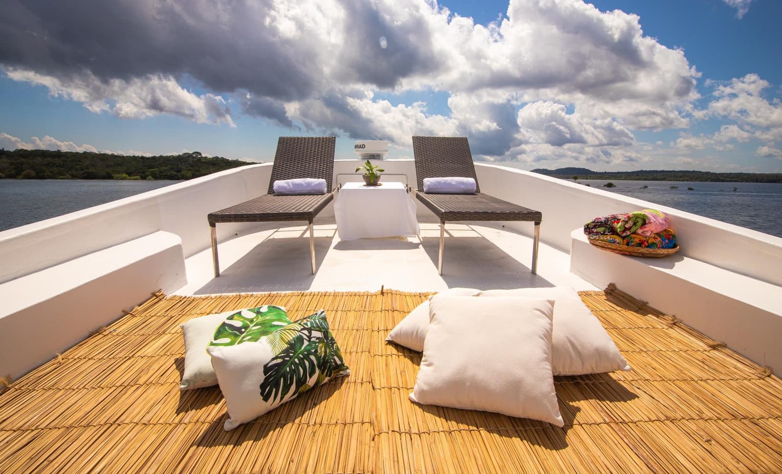Das Büro für heute Nachmittag, Ausblick inklusive, Dresscode optional. Zwei Korbliegen mit gerollten weißen Handtüchern auf dem offenen Bugdeck einer Amazonas-Kreuzfahrt ab Alter do Chão, mit tropischen Bodenkissen, einer Bambusmatte und bewaldeten Flussufern unter einem dramatisch bewölkten Himmel