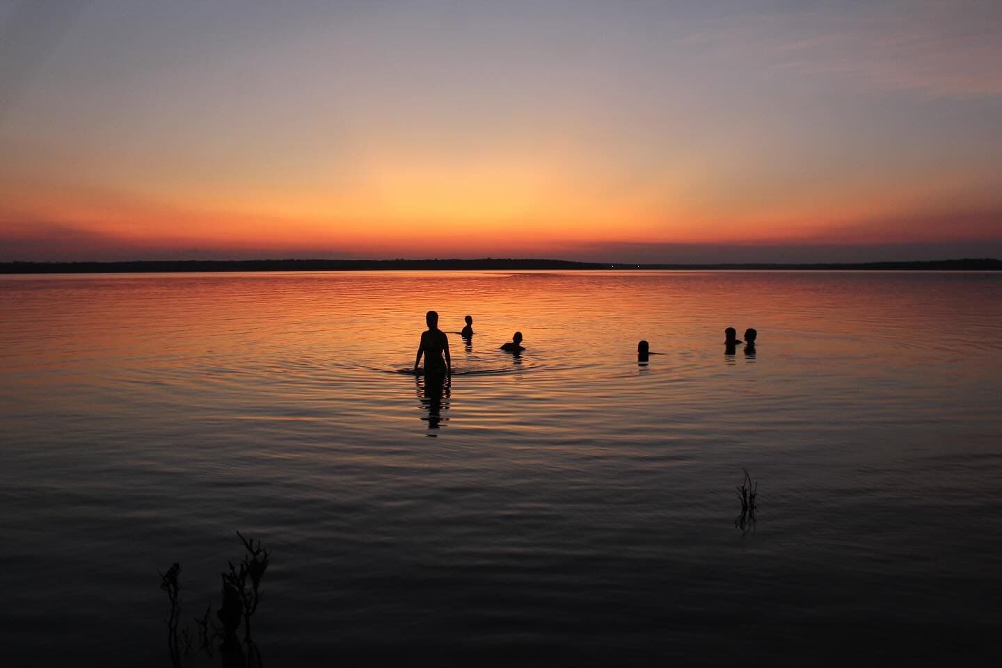 Der Fluss hält den Tag fest, solange man ihn lässt. Silhouetten von Reisenden, die in einem weiten, spiegelglatten Fluss in der Abenddämmerung schwimmen, während ein feuriger Orange-Rot-Sonnenuntergang sich im Wasser spiegelt – auf einer Amazon-Kreuzfahrt ab Alter do Chão