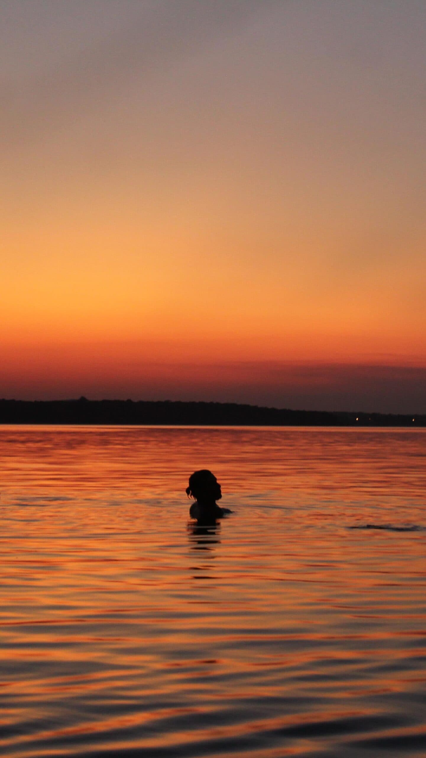 Manche Abende lassen sich am besten im Wasser erleben. Eine Person schwimmt bei Sonnenuntergang in einem weiten Fluss, als dunkle Silhouette vor einem tief orangefarbenen Abendhimmel auf einer Amazon-Kreuzfahrt ab Alter do Chão