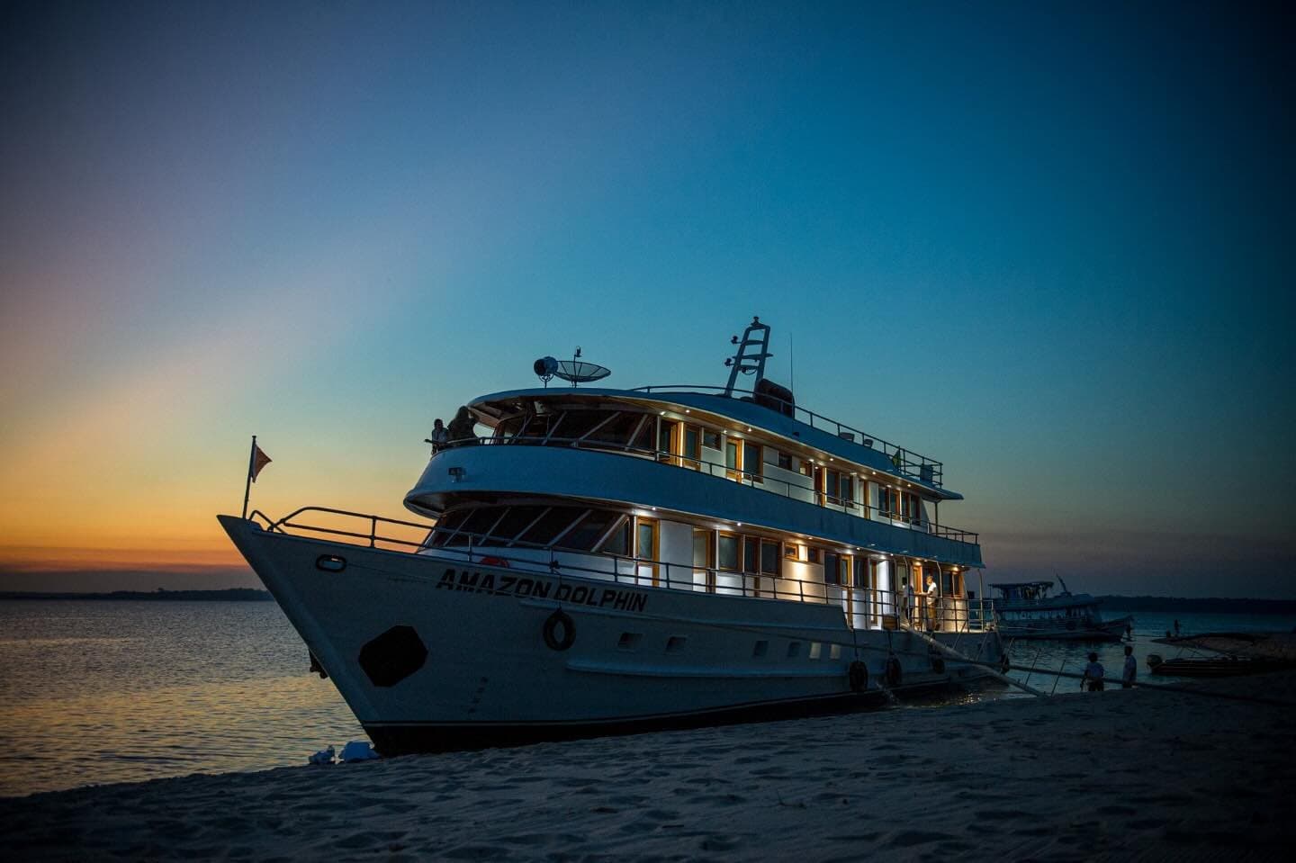 Das schwimmende Lager wartet, bevor der Fluss in die Nacht taucht. Das Kreuzfahrtschiff Amazon Dolphin liegt bei Einbruch der Dämmerung an einer Sandbank, die warmen Kabinenlichter leuchten vor einem tiefblauen und orangefarbenen Abendhimmel