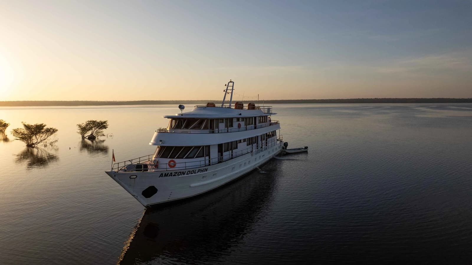 Hier draußen auf dem Fluss beginnt der Abend ganz langsam zu leuchten. Das Flussschiff Amazon Dolphin liegt im goldenen Abendlicht auf einem spiegelglatten Amazonas-Nebenfluss vor Anker, umgeben von überfluteten Uferbäumen
