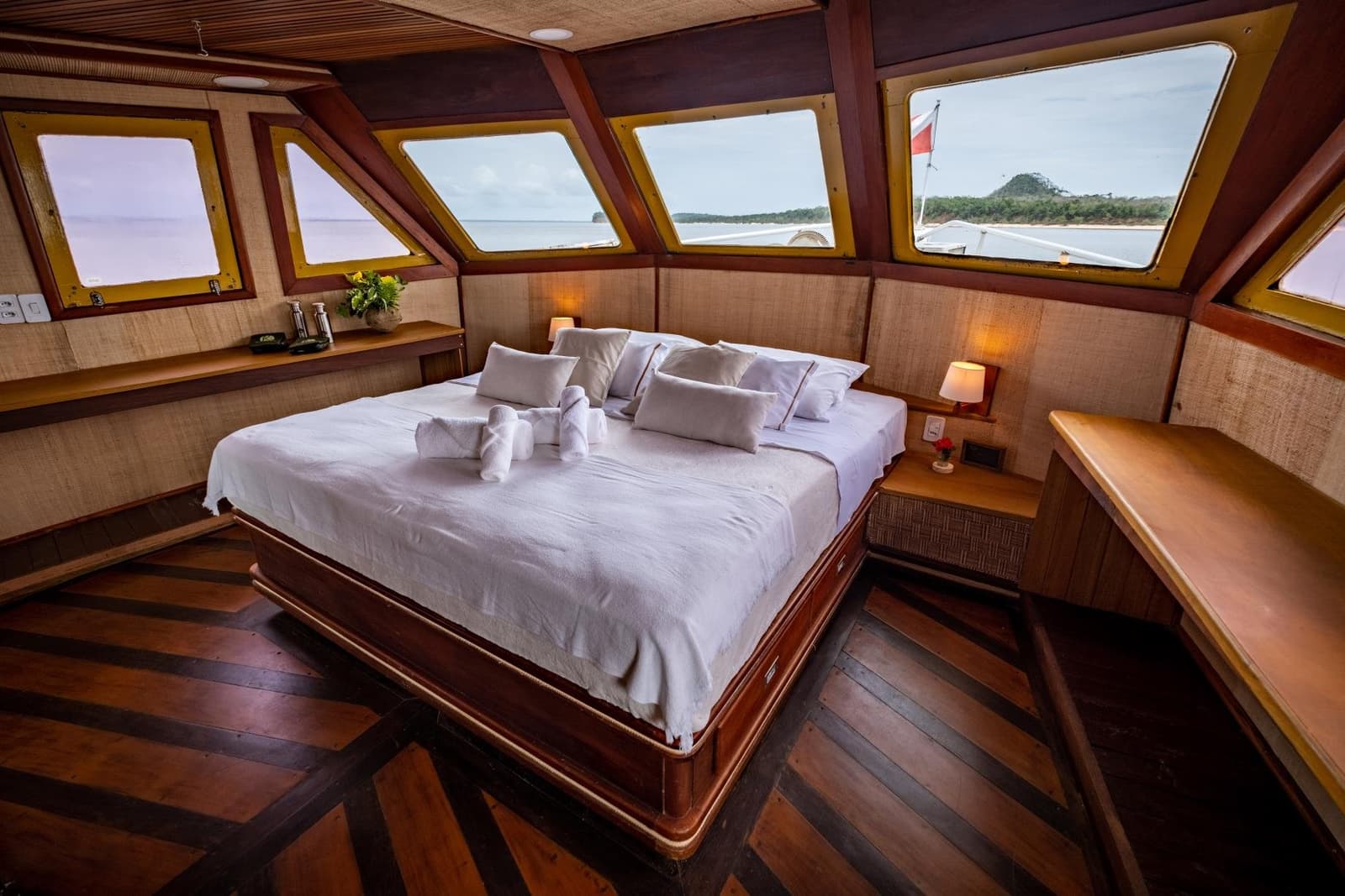 Jeden Morgen ein neues Panorama, direkt vom Bett aus. Geräumige Hauptkabine auf einer Amazonas-Kreuzfahrt ab Alter do Chão mit Kingsize-Bett in weißer Bettwäsche, gefalteten Handtuchtieren, warmem Holzdekor und Panoramafenstern mit Blick auf ein bewaldetes Flussufer