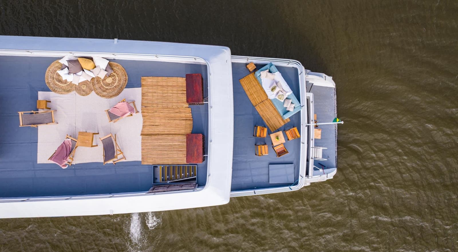 Ein Wohnzimmer mit Aussicht, das sich langsam flussaufwärts bewegt. Luftaufnahme eines Amazon-Kreuzfahrtschiffs ab Alter do Chão mit zwei möblierten Sonnendecks: Holzliegen, Rattanteppiche, Strohdächer und gepolsterte Sitzgruppen auf breitem braunen Fluss