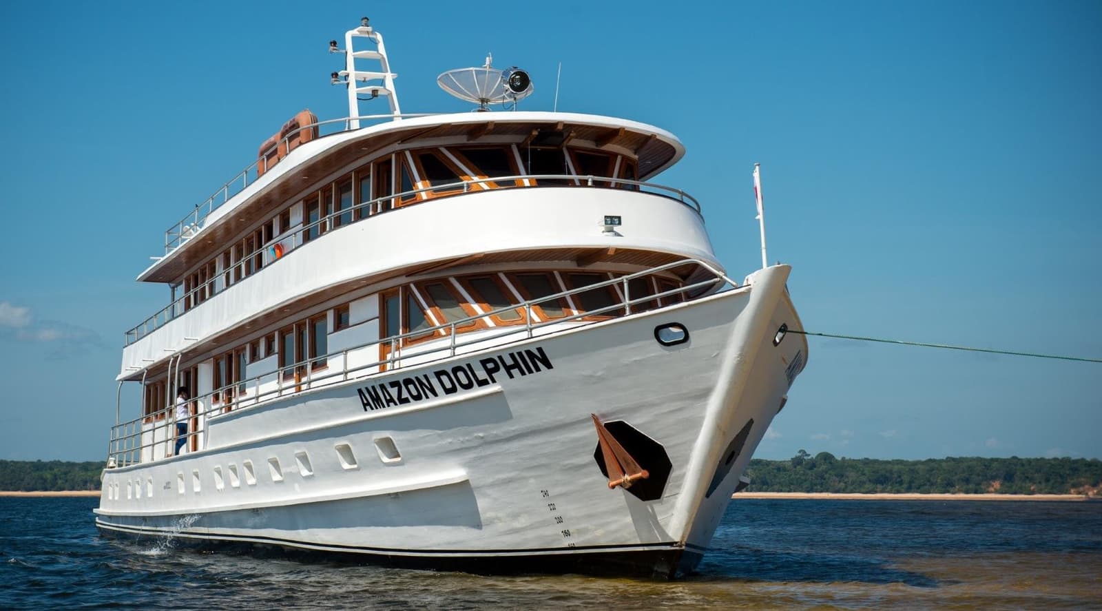 Drei Decks, ein Fluss, kein WLAN-Stress, so soll Urlaub sein. Das mehrstöckige Flusskreuzfahrtschiff Amazon Dolphin mit weißem Rumpf und warmem Teakholz-Dekor fährt auf einem breiten Flusskanal unter blauem Himmel — Teil der Amazon-Kreuzfahrt ab Alter do Chão