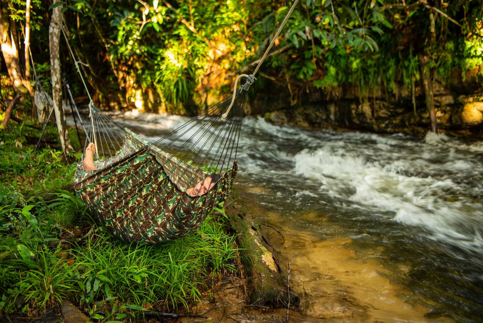 Der Fluss rauscht, die Hängematte schaukelt, die Zeit steht still. Ein Reisender ruht in einer Tarnmuster-Hängematte, die neben einem rauschenden Dschungelbach zwischen tropischen Bäumen aufgehängt ist, auf einer Amazonas-Kajakexpedition in Brasilien
