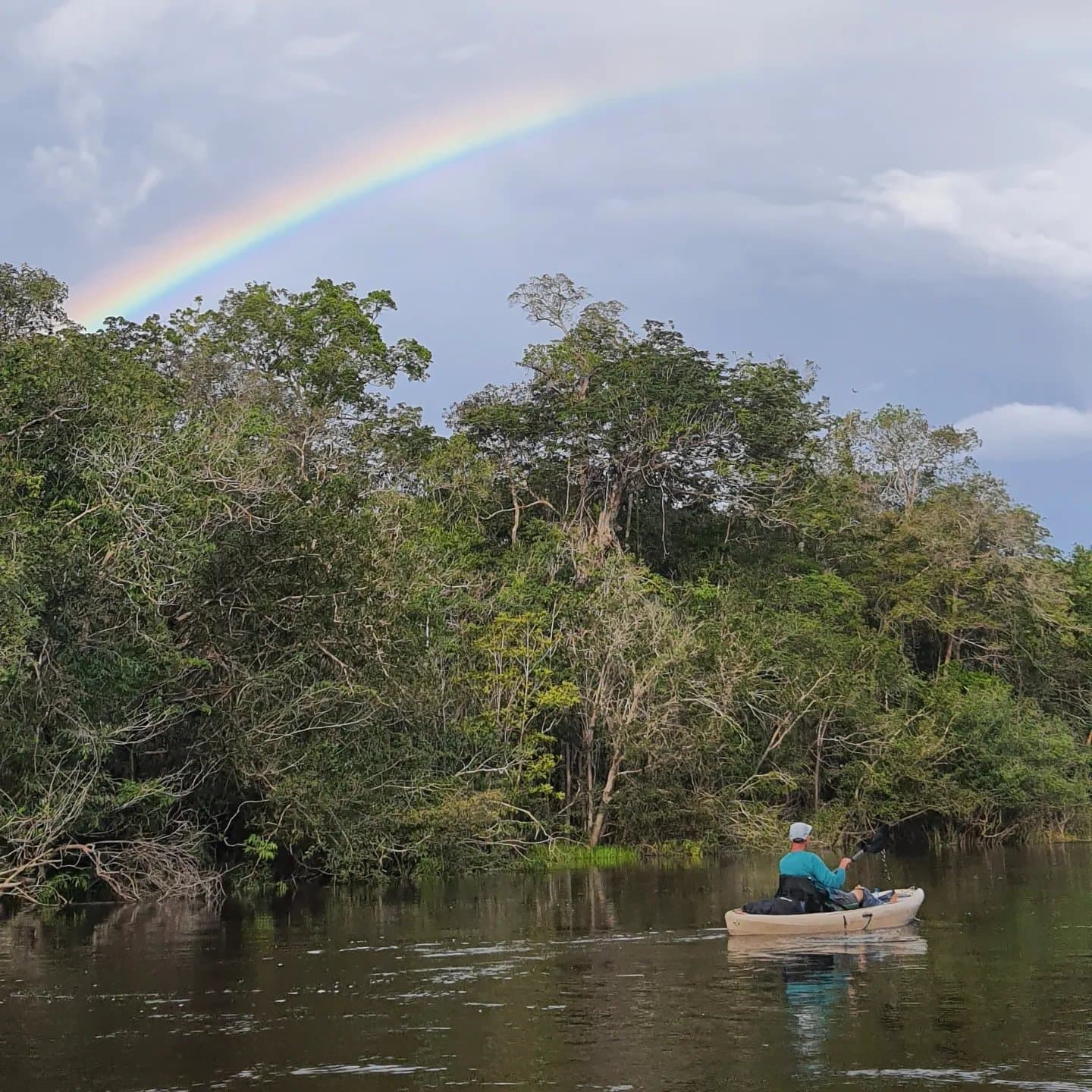 Manche Paddeltouren enden mit einem Regenbogen. Diese hier auch. Ein Kajakfahrer paddelt auf einem dunkelwasserigen Fluss in Richtung dichtem Regenwald, während ein leuchtender Regenbogen über dem bewölkten Himmel eines Amazonas-Kajak-Abenteuers erscheint