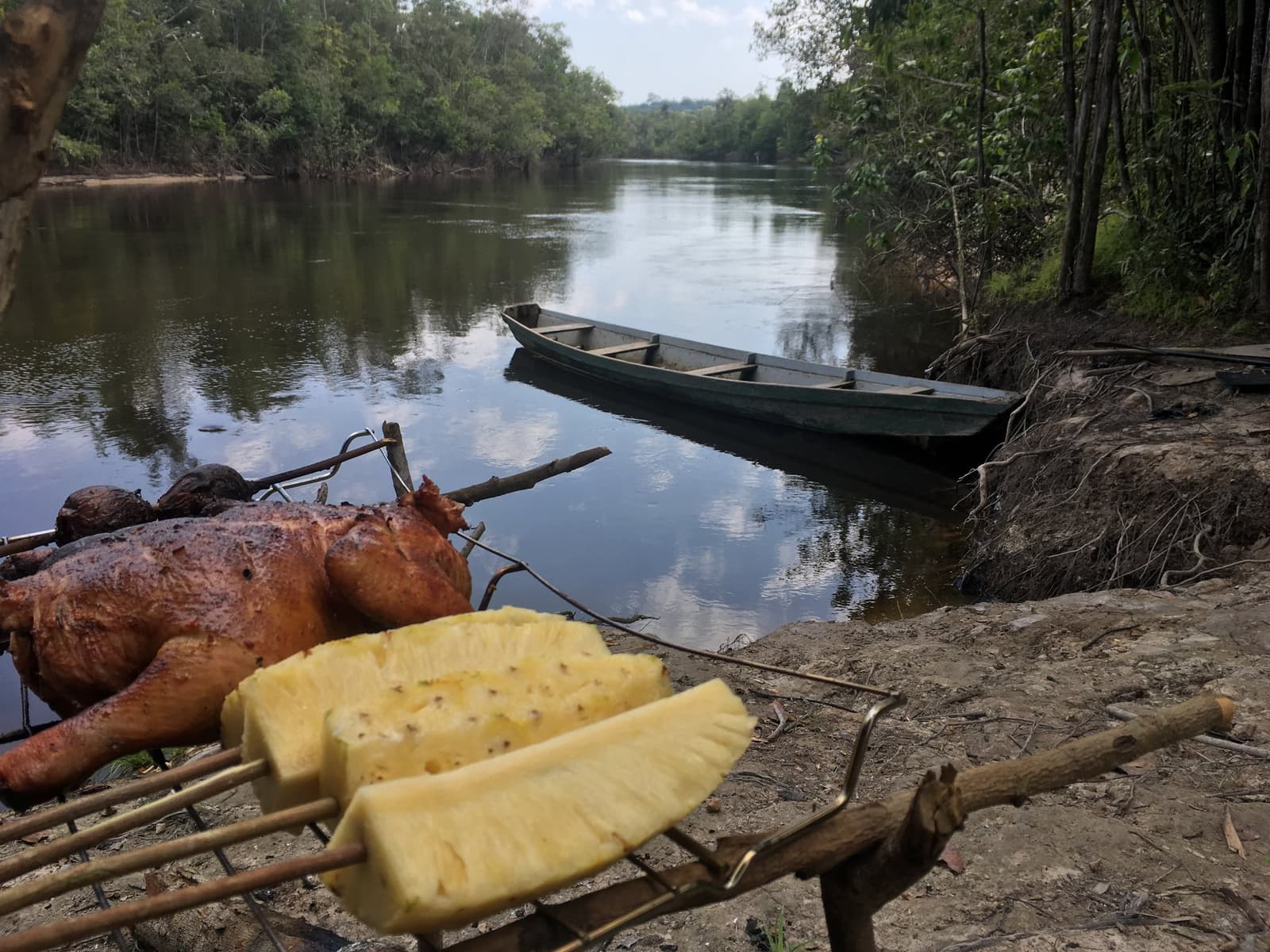 Mittagessen am Ufer, Kanu wartet, hier hat der Amazonas das letzte Wort. Gegrilltes Fleisch und frische Ananasscheiben auf Bambusspießen am Flussufer beim Amazon-Kajaktouren in Brasilien, mit einem Holzkanu auf einem ruhigen Dschungelfluss im Hintergrund