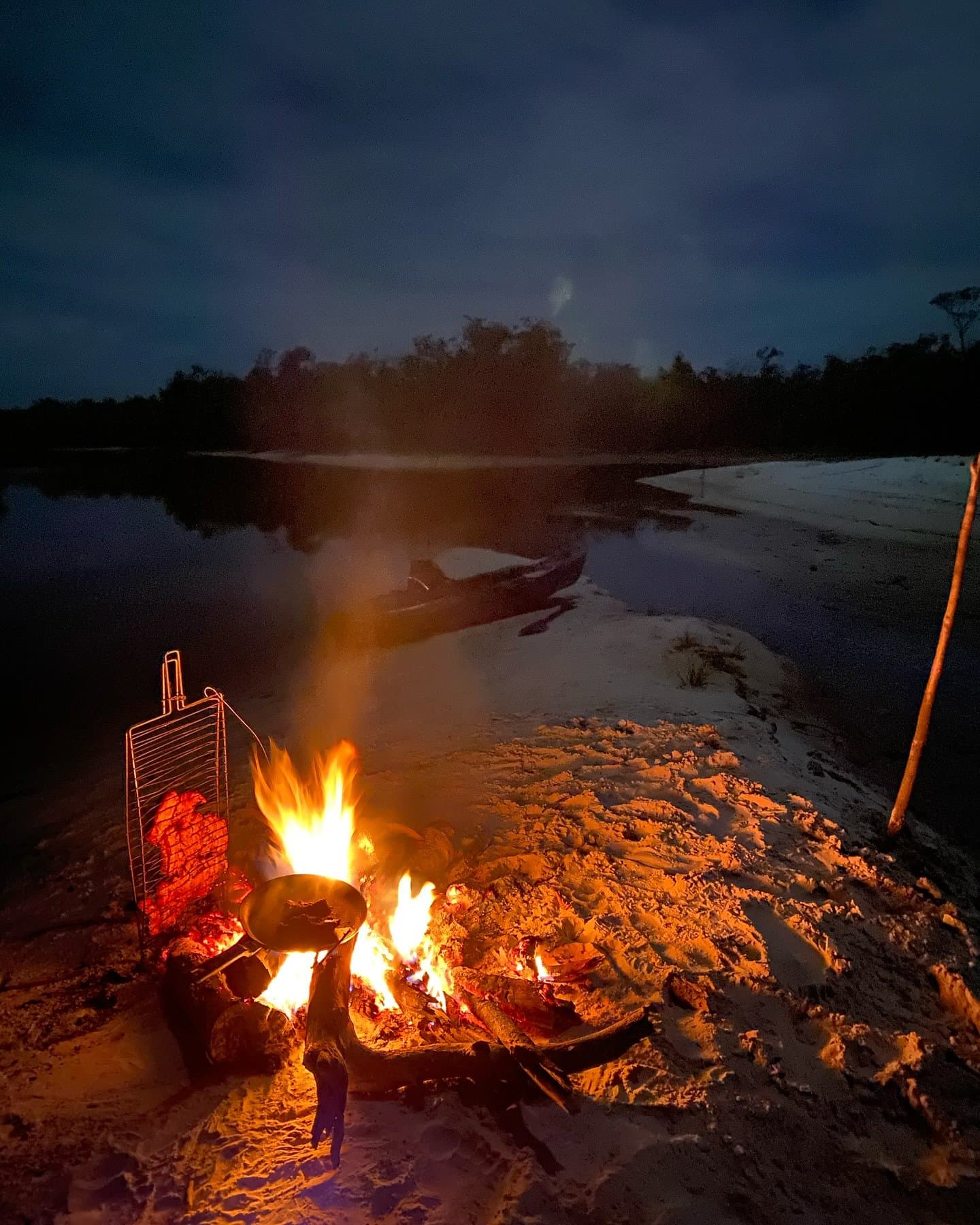 Feuer an, Boot vertäut, das Abendessen kocht sich fast von selbst. Ein knisterndes Lagerfeuer mit Kochpfanne und Grillrost an einem weißen Sandstrand am Fluss in der Nacht, mit einem Holzkanu am Ufer während einer Amazonas-Kajaktour in Brasilien