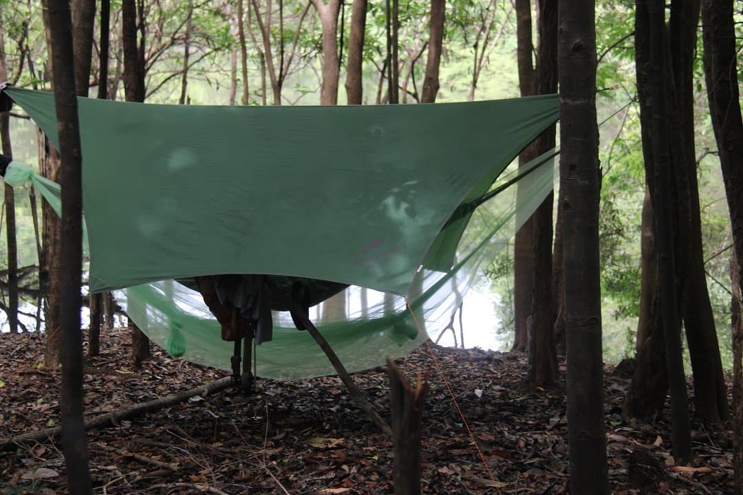 Kein Dach, kein Lärm, kein Problem, so schläft man im Amazonas. Grüne Plane zwischen Bäumen gespannt über einer Hängematte mit Moskitonetz auf einer Amazon-Survival-Tour, Laubstreu am Waldboden und Wasser im Hintergrund