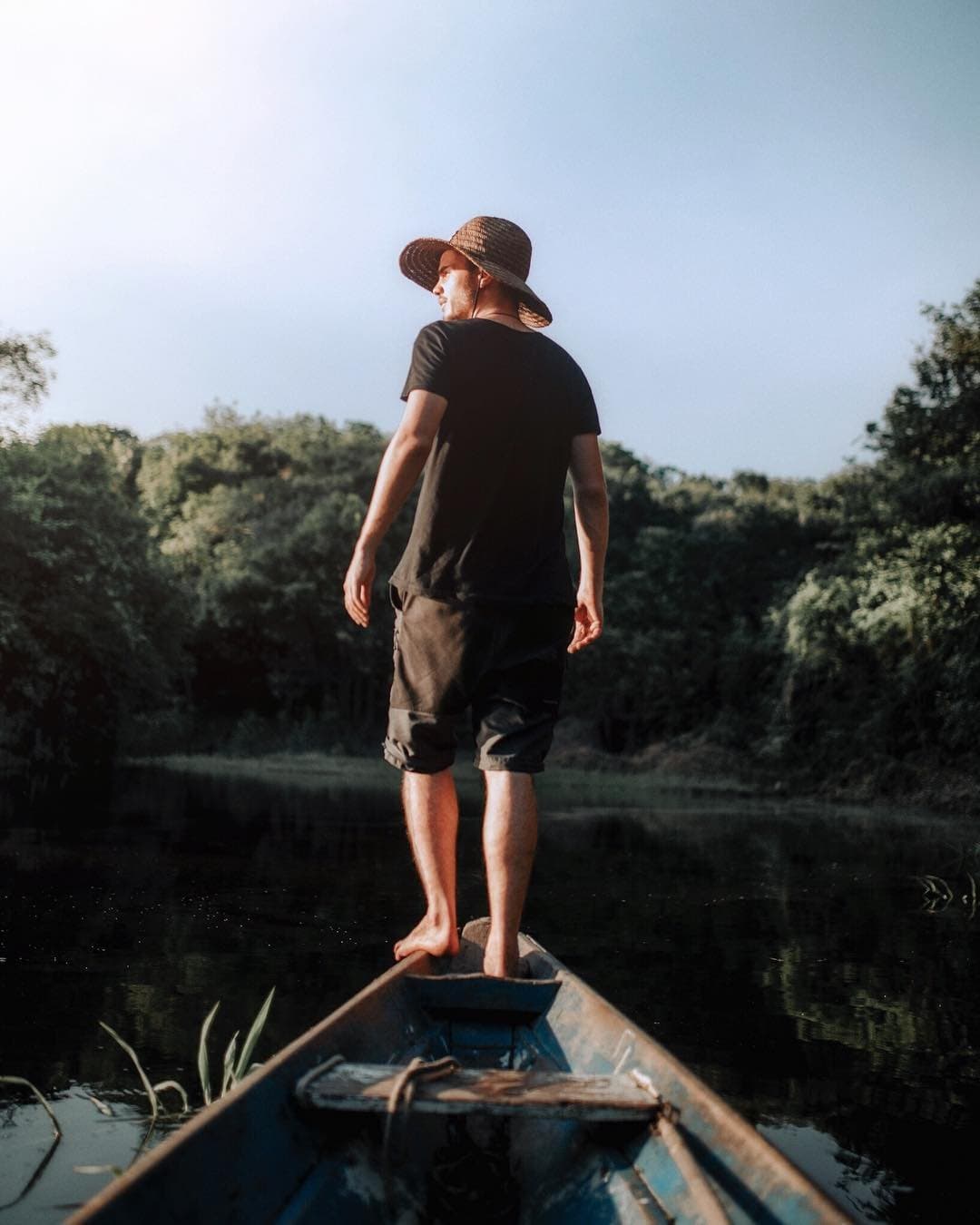 Pieds nus sur la proue, le fleuve décide de la suite. Un homme pieds nus coiffé d'un chapeau de paille se tient à la proue d'une vieille pirogue en bois sur une rivière sombre et calme bordée d'une forêt dense