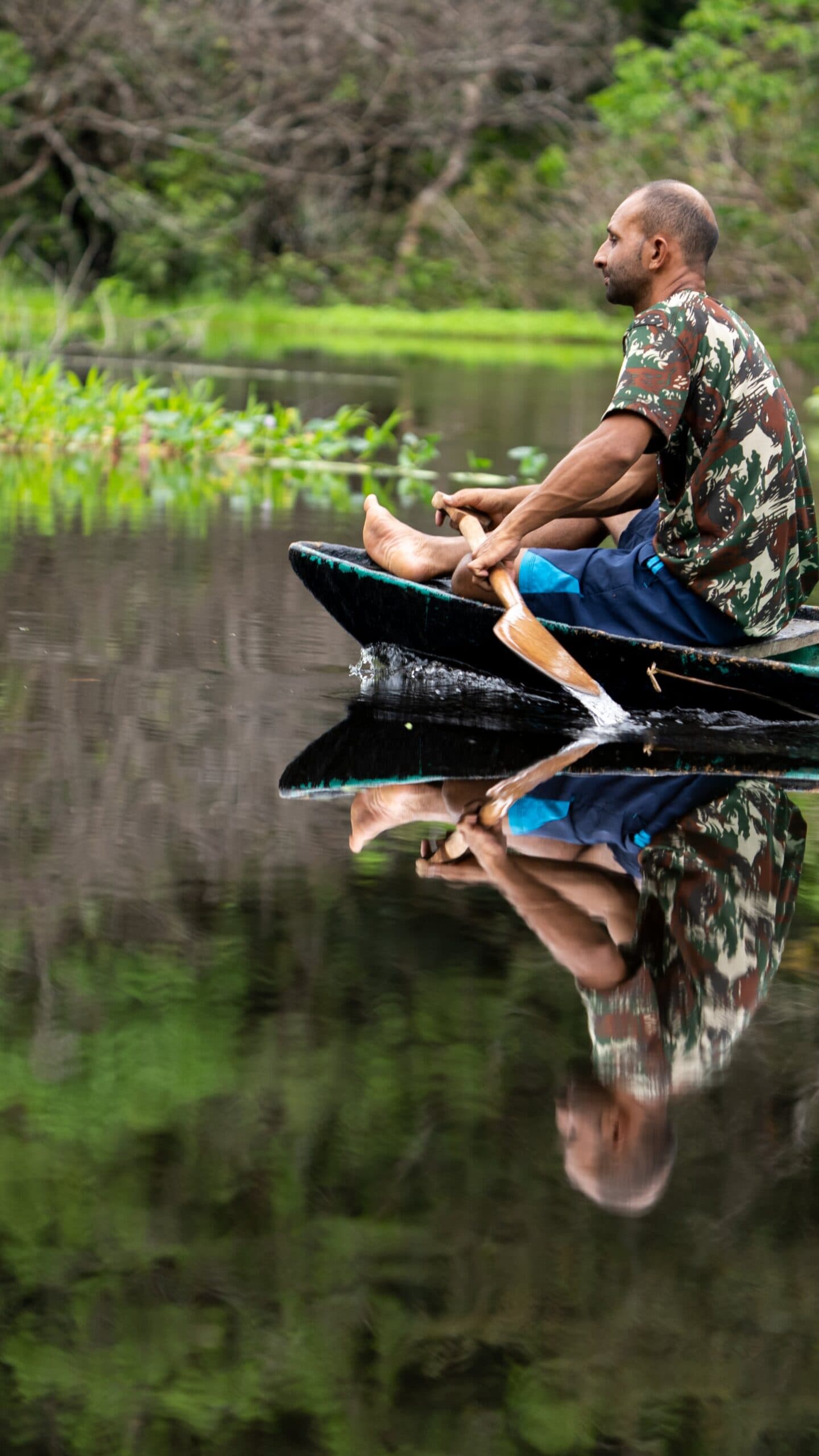 Pas de GPS, pas besoin. Il lit le fleuve mieux que personne. Un homme pagaie un petit canoë en bois sur un cours d'eau amazonien aux eaux noires et lisses comme un miroir, son reflet parfait visible à la surface immobile, avec une végétation tropicale luxuriante en arrière-plan — un instant suspendu lors d'un trek de survie en Amazonie