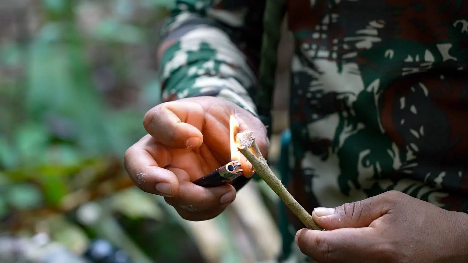 Un geste précis, une flamme, et la forêt qui regarde. Un guide en tenue camouflage allume un bâton vert à l'aide d'un briquet en forêt tropicale, démontrant une technique de survie en pleine jungle lors d'un trek et tour de survie en Amazonie