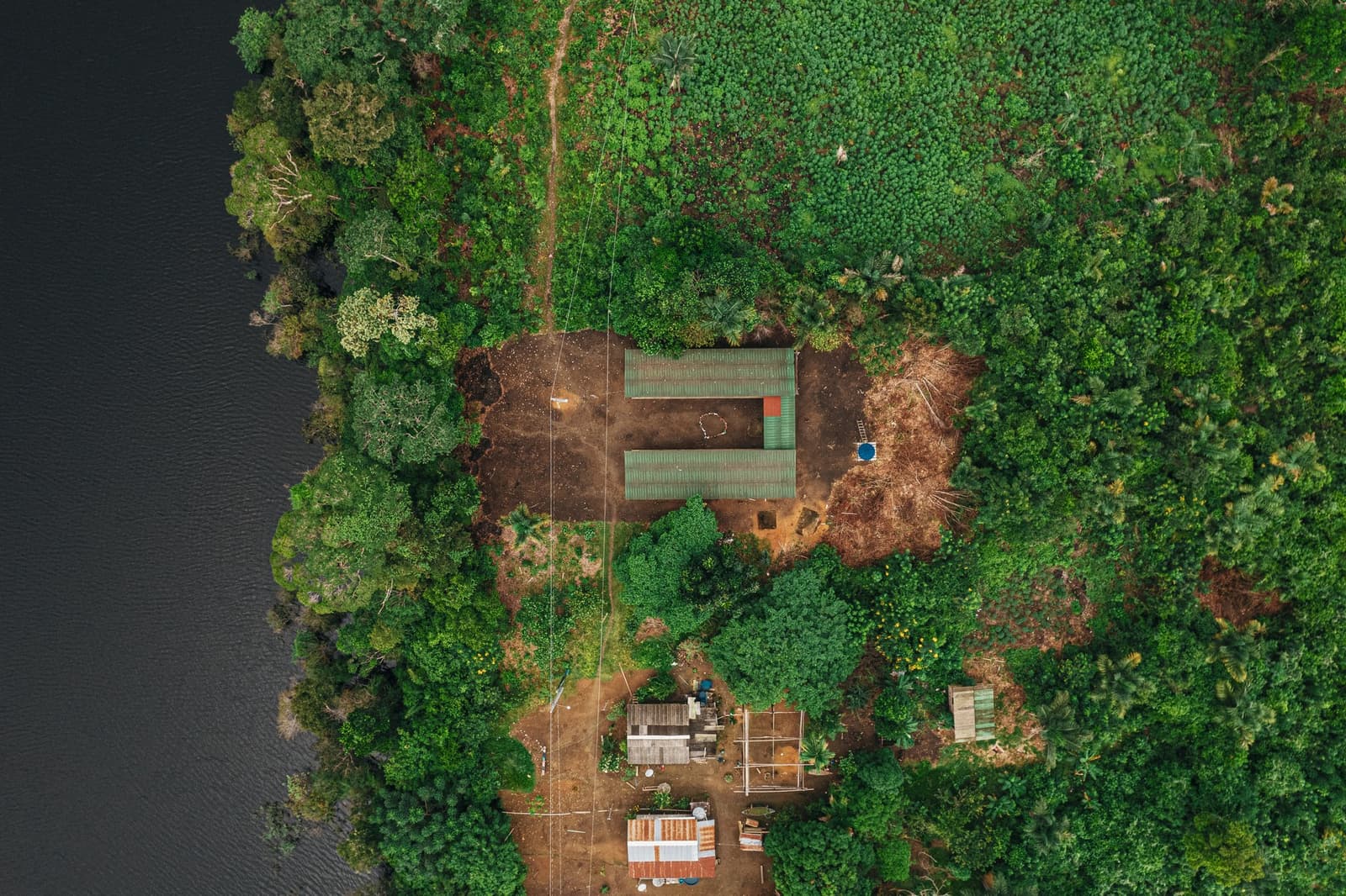 Hier holt sich der Dschungel alles zurück, was er für einen Moment hergibt. Drohnenaufnahme aus der Vogelperspektive der Anavilhanas Jungle Lodge Brasilien, mit grünen Wellblechdächern inmitten dichtem Regenwalddach neben einem dunklen Fluss und einer kleinen Ufergemeinschaft