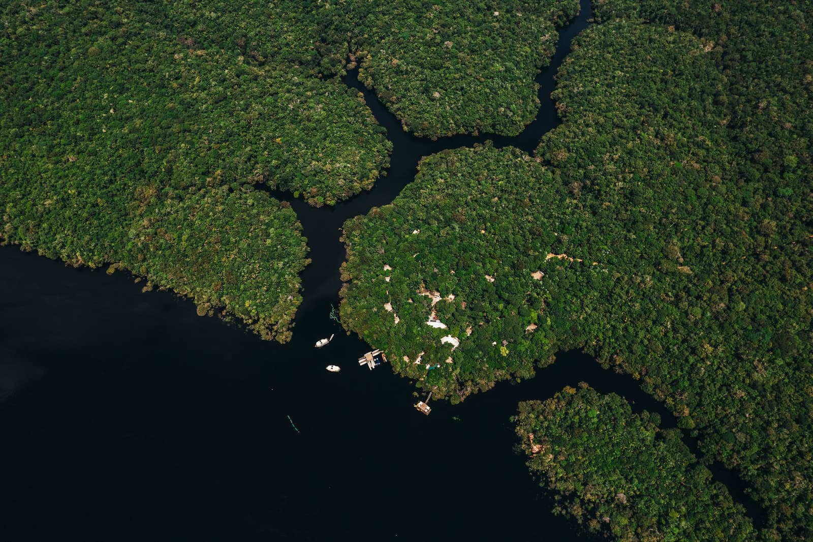 Winzige Boote, endloser Wald, das Lodge weiß genau, wo es hingehört. Luftaufnahme der Anavilhanas Jungle Lodge Brasilien mit kleinen Booten und einem Schwimmdock am Rand einer dichten Regenwaldsinsel inmitten dunkler Wasserarme des Rio Negro