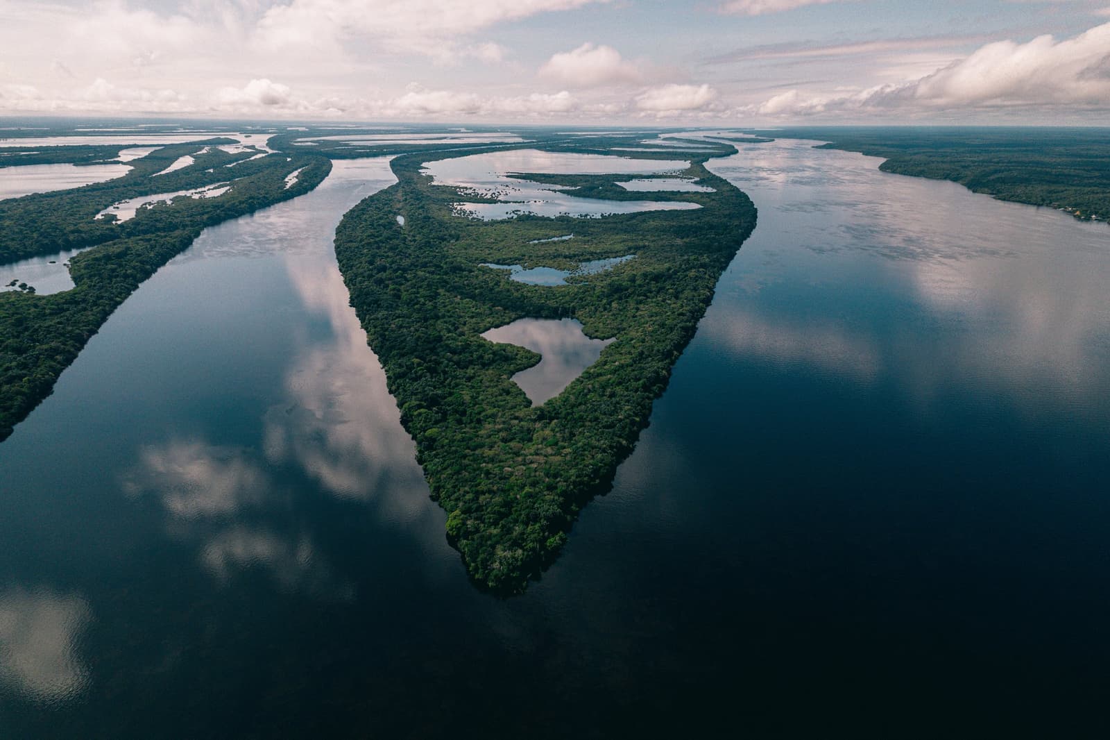 So viele Inseln, dass der Fluss selbst den Überblick verliert. Luftaufnahme einer dicht bewaldeten Insel im Anavilhanas Jungle Lodge Brazil, umgeben von den dunkel-spiegelnden Gewässern eines weitläufigen Flussarchipels unter bewölktem Himmel