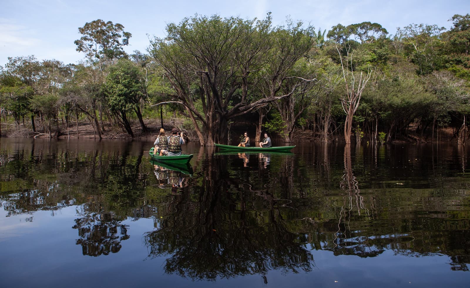 Hier hat man das Gefühl, der Fluss atmet ganz langsam. Drei Reisende in einem grünen Kanu gleiten durch einen überschwemmten Wald auf spiegelglattem dunklem Wasser in der Anavilhanas Jungle Lodge Brasilien, umgeben von halb versunkenen Bäumen und ihren perfekten Spiegelbildern