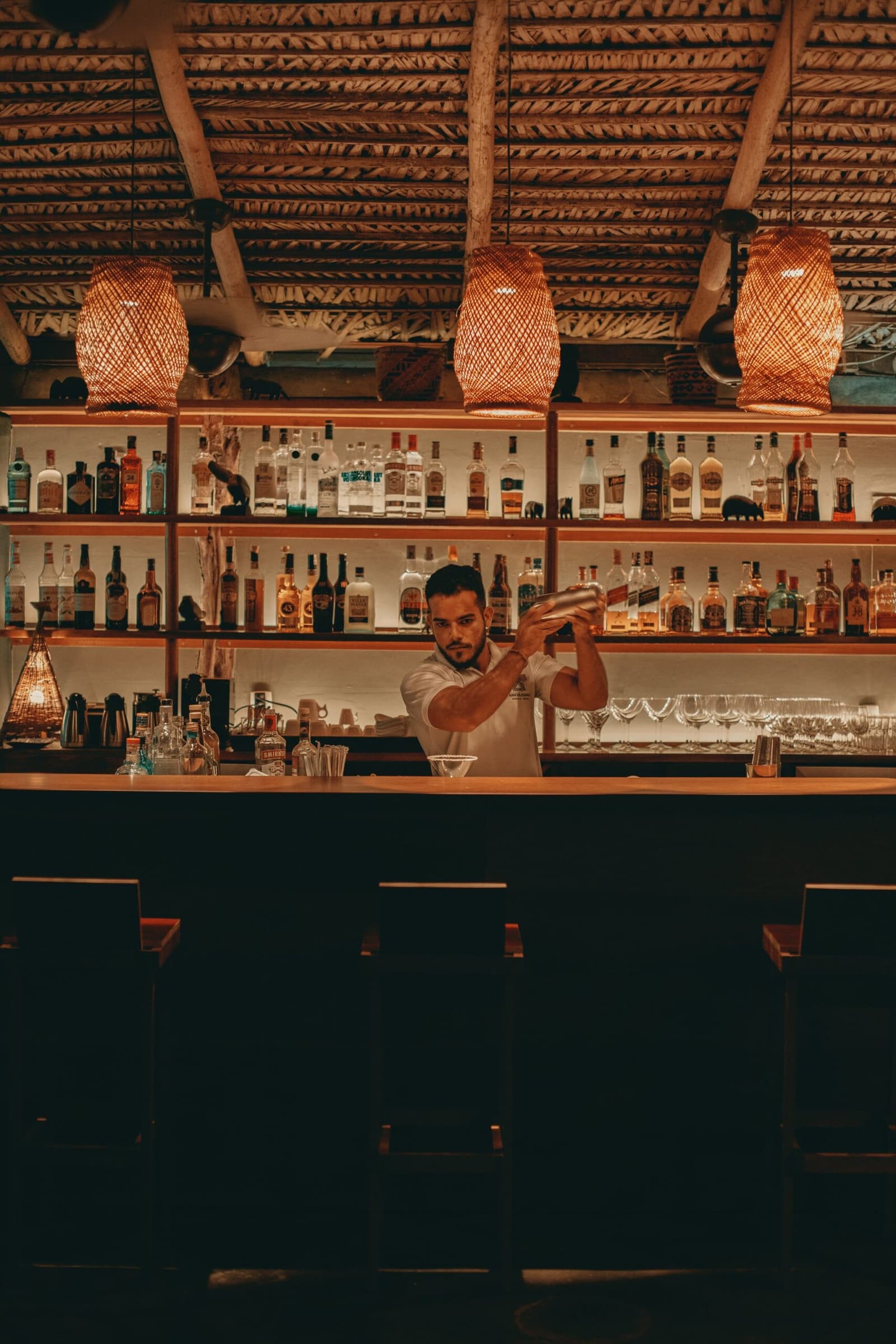 Der Dschungeltag klingt aus, und die Bar ist längst bereit. Ein Barkeeper schüttelt einen Cocktail-Shaker hinter der Bar der Anavilhanas Jungle Lodge Brazil, beleuchtet von warmem Rattanlicht und umgeben von Spirituosenregalen