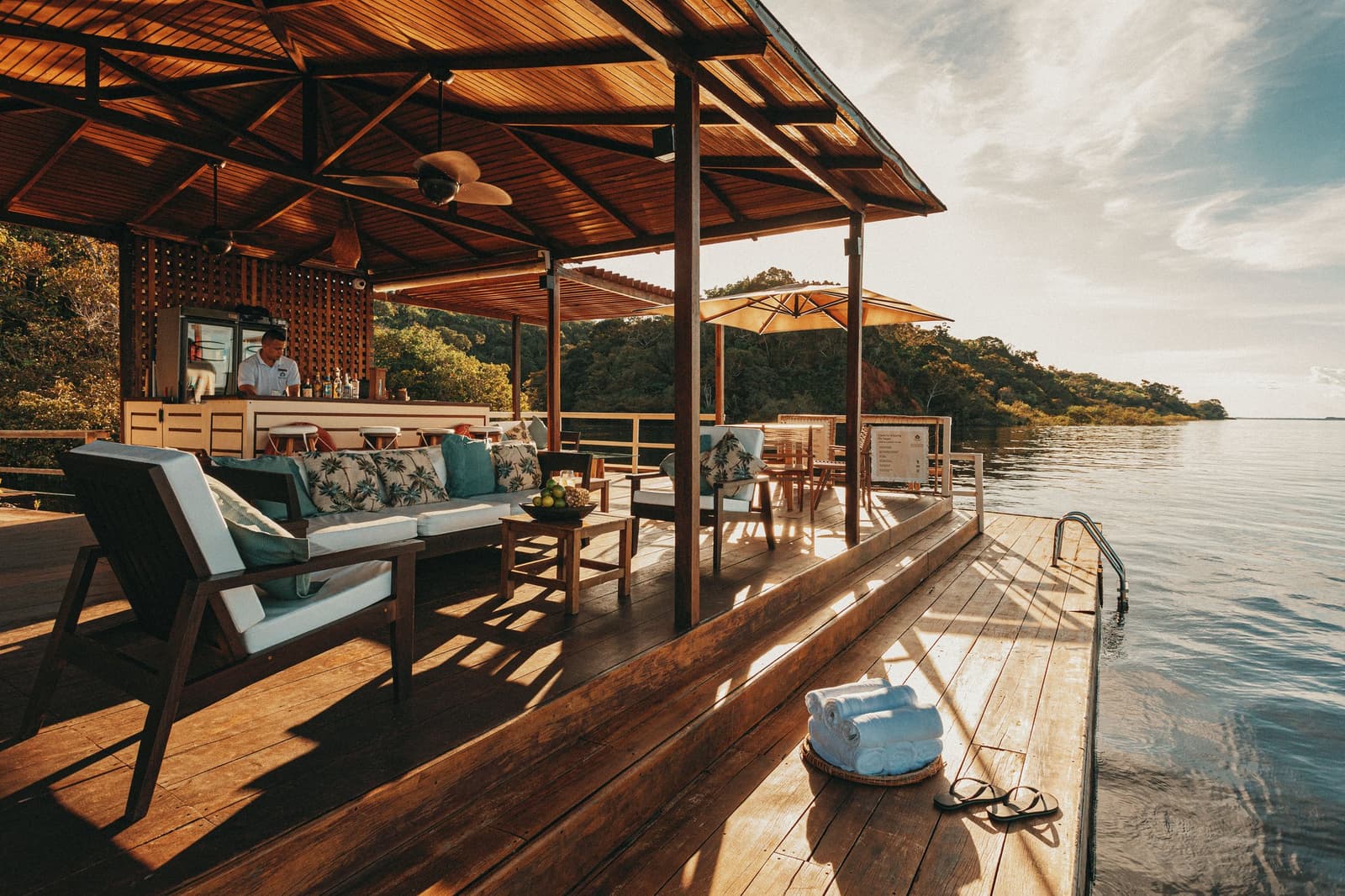 Ein Sundowner schmeckt besser, wenn der Amazonas direkt vor der Terrasse liegt. Holzdeck der Anavilhanas Jungle Lodge in Brasilien mit Lounge-Sofas, einem besetzten Bartresen und gefalteten Handtüchern am Rand des ruhigen Rio Negro, im Hintergrund bewaldete Inseln