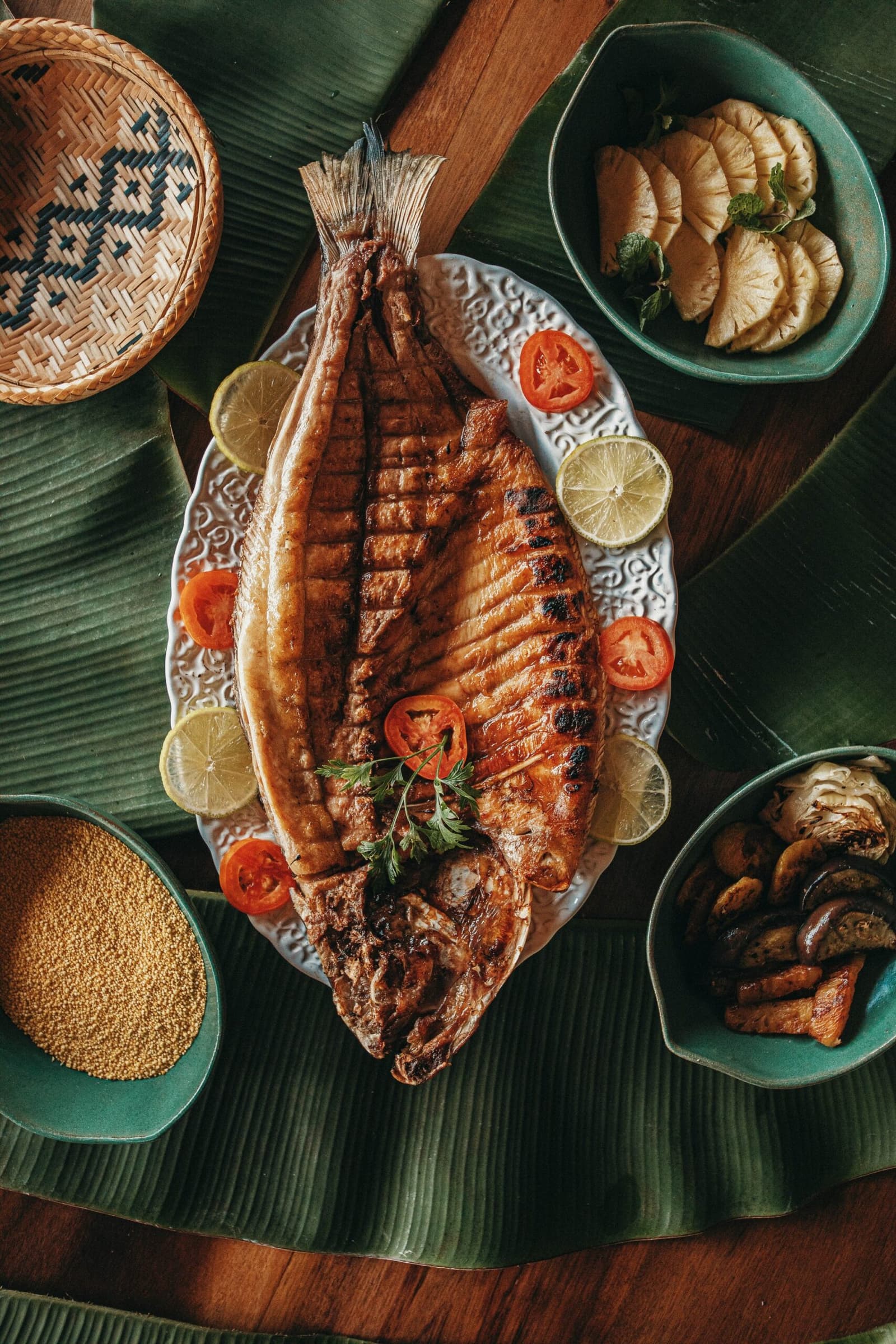 Frisch aus dem Fluss, direkt auf den Tisch, besser geht es kaum. Ein ganzer gegrillter Fisch auf einer verzierten weißen Platte mit Limetten- und Tomatenscheiben in der Anavilhanas Jungle Lodge Brasilien, umgeben von Beilagen mit Ananas, gegrillten Pilzen und goldenen Samen auf Bananenblättern