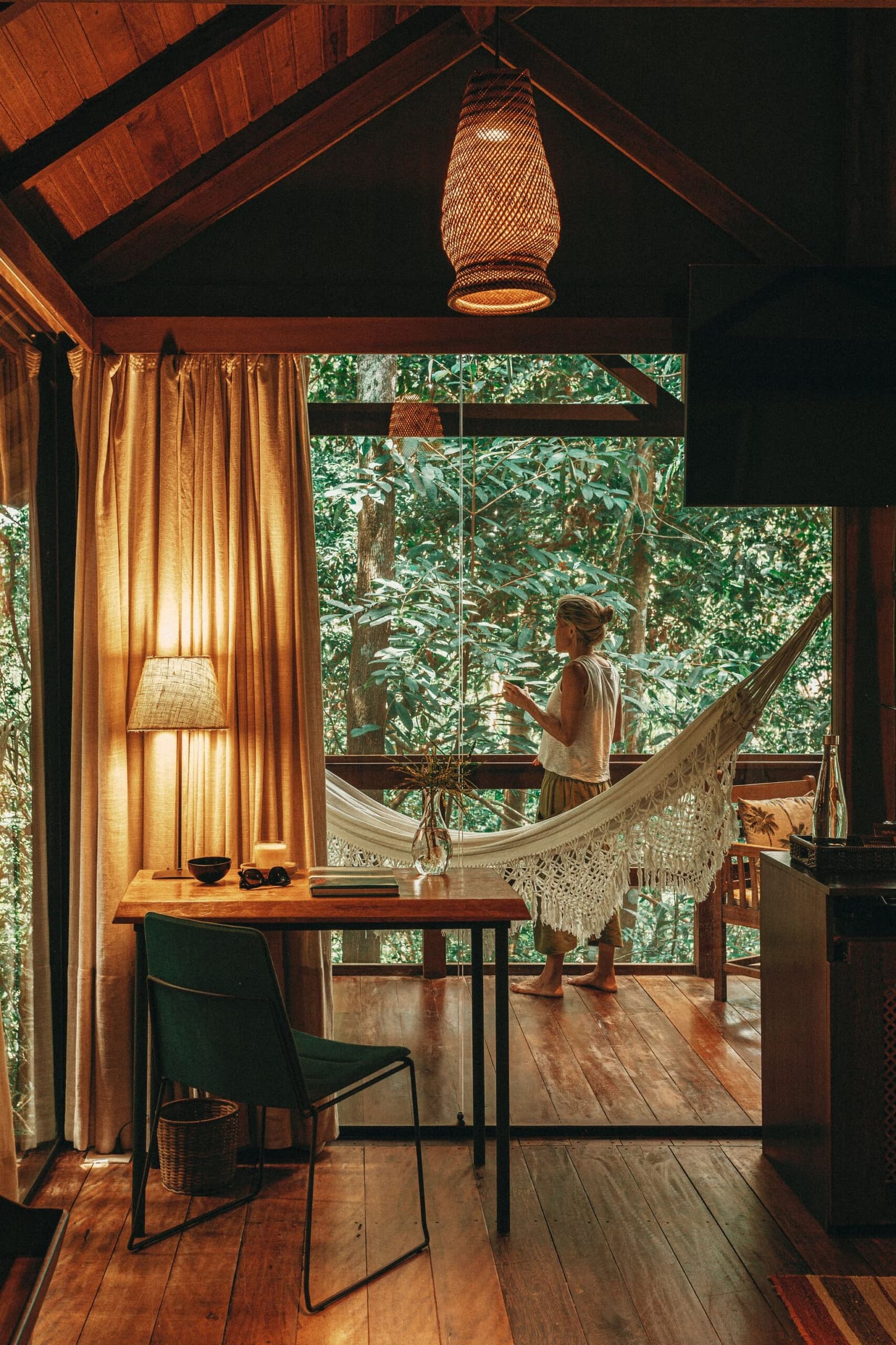 Kaffee, Urwald, Hängematte. Manche Morgen brauchen keinen Plan. Innenraum einer Holzhütte im Anavilhanas Jungle Lodge Brazil mit warmem Holzboden, einem geflochtenen Hängelampe und einer Frau, die barfuß auf einer mit Hängematte ausgestatteten Veranda in den dichten Regenwald blickt