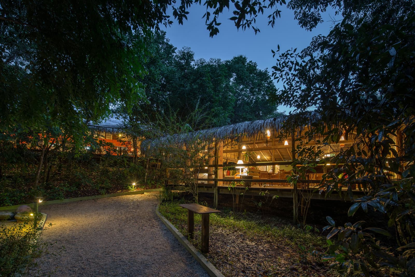 Wenn die Dämmerung kommt, weist das warme Licht der Lodge den Weg nach Hause. Ein mit Poller-Lichtern gesäumter Kiesweg führt durch dichten Tropenwald zur beleuchteten Tiki-Pavillon-Terrasse der Anavilhanas Jungle Lodge Brazil in der Abenddämmerung