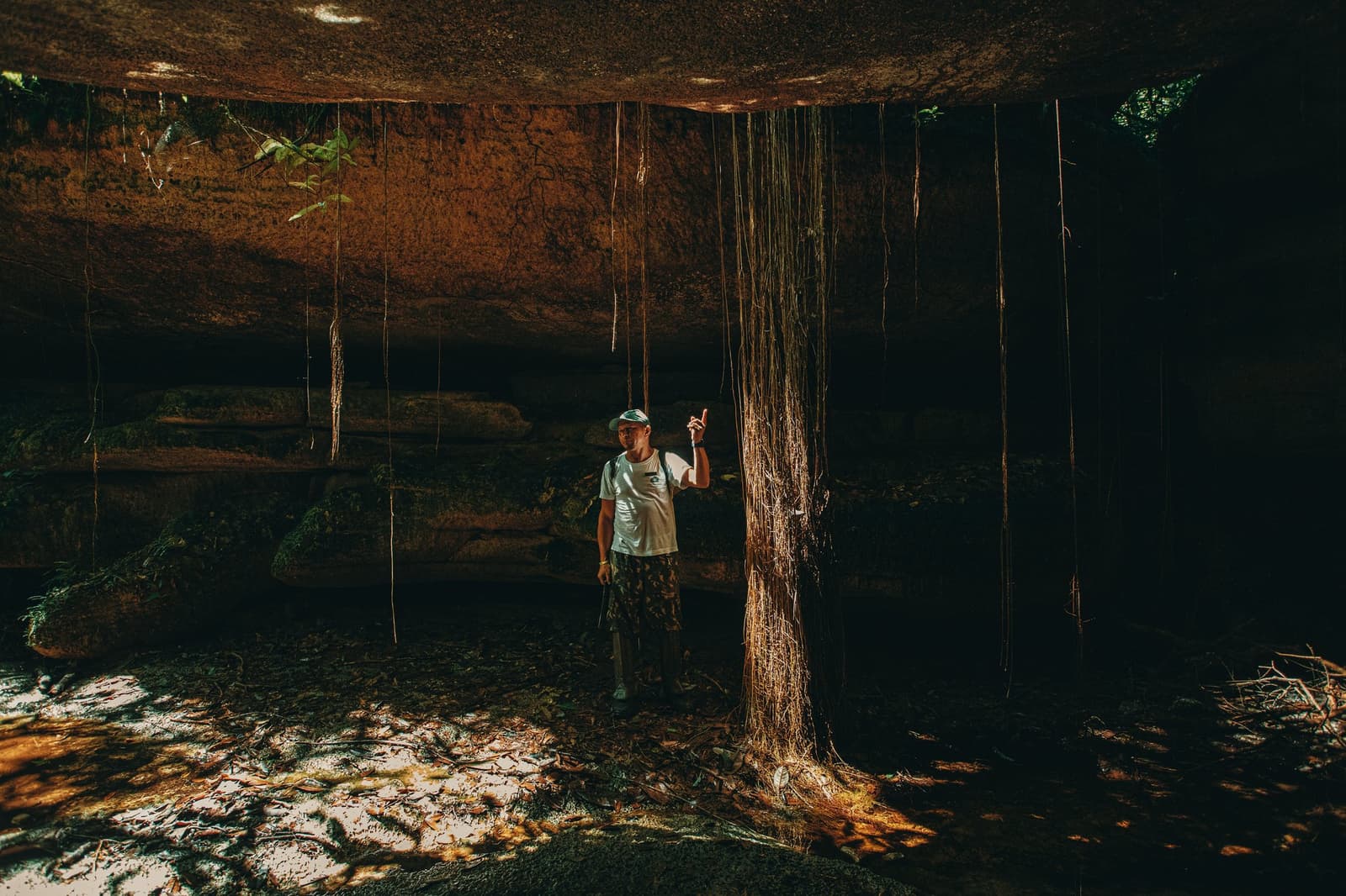 Selbst unter der Erde hat der Regenwald noch Geschichten zu erzählen. Ein Führer zeigt nach oben unter einem massiven uralten Felsüberhang in den Madadá-Höhlen, Anavilhanas Jungle Lodge Brasilien, mit langen Luftwurzeln, die in dramatischem goldenen Licht von der Decke hängen