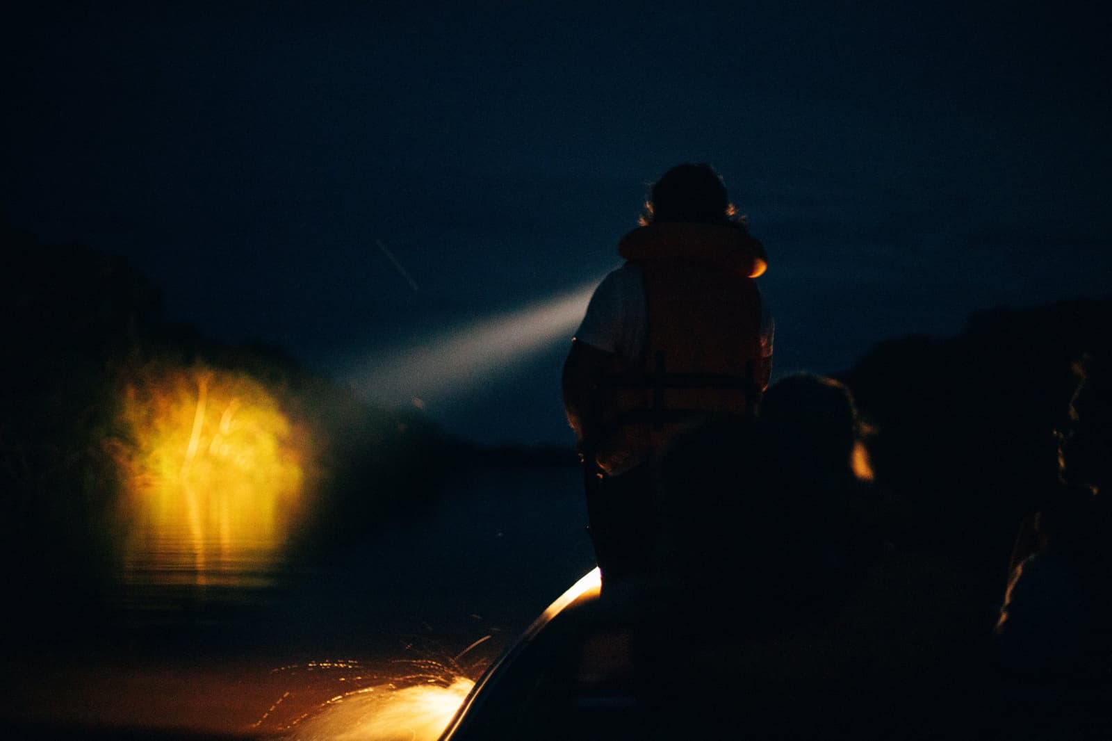 Der Fluss gibt nicht viel preis. Der Scheinwerfer versucht es trotzdem. Ein silhouettierter Guide in einer Schwimmweste leuchtet mit einem Suchscheinwerfer über dunkles Flusswasser bei einer Nachttour in der Anavilhanas Jungle Lodge Brazil, während vorne ein warmgelbes Licht glüht