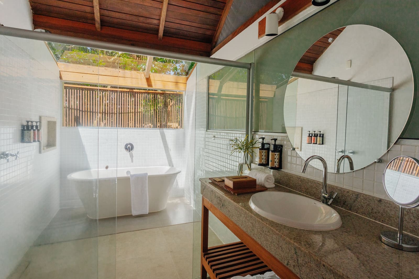 Ein Bad mit Waldblick, weil Komfort und Regenwald sich nicht ausschließen. Offenes Badezimmer der Anavilhanas Jungle Lodge Brazil mit freistehender Badewanne unter einem Holzlamellenschirm, Granitwaschbecken, Rundspiegel und Holzbalkendecke mit Blick in die Baumkronen