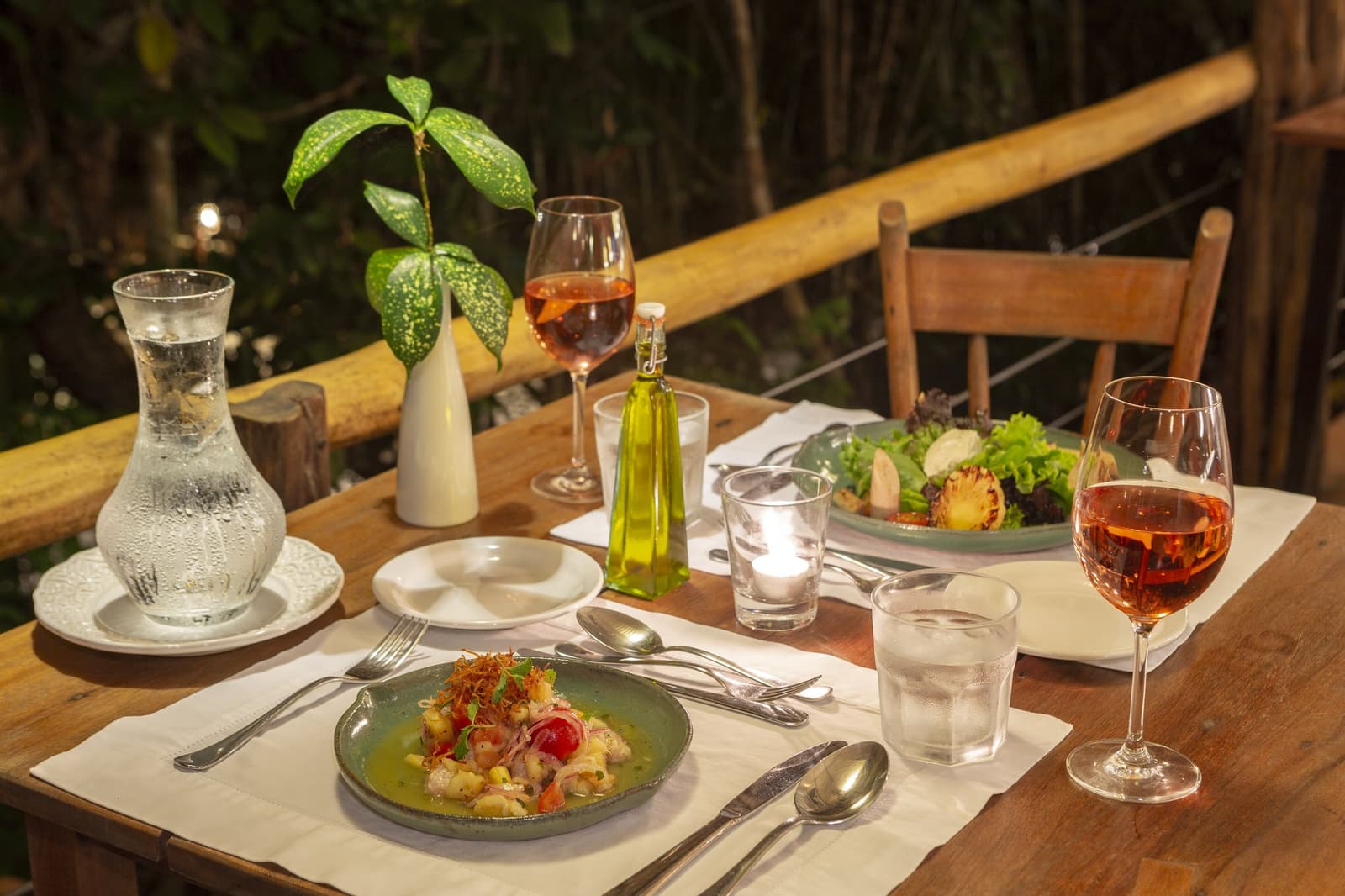 Frisch, lokal, und mit Blick auf den Regenwald, so schmeckt der Amazonas. Kerzenschein-Abendtisch im Anavilhanas Jungle Lodge Brazil mit Roséwein, einem grünen Keramikteller mit Meeresfrüchte-Vorspeise, frischem Salat und einem tropischen Blatt-Mittelstück auf einer Bambusterrasse mit Blick in den Dschungel