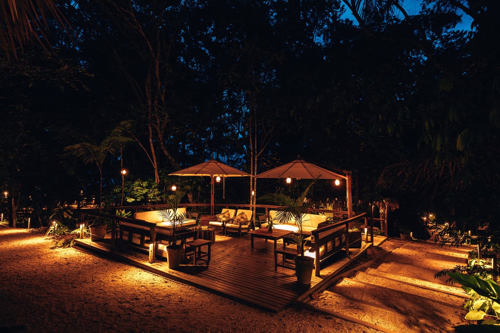 Abende hier verlaufen ohne Eile, der Regenwald gibt den Takt vor. Beleuchtete Open-Air-Terrasse der Anavilhanas Jungle Lodge Brazil mit gepolsterten Holzsofas unter Sonnenschirmen und warmem Laternenlicht im Regenwald bei Dämmerung