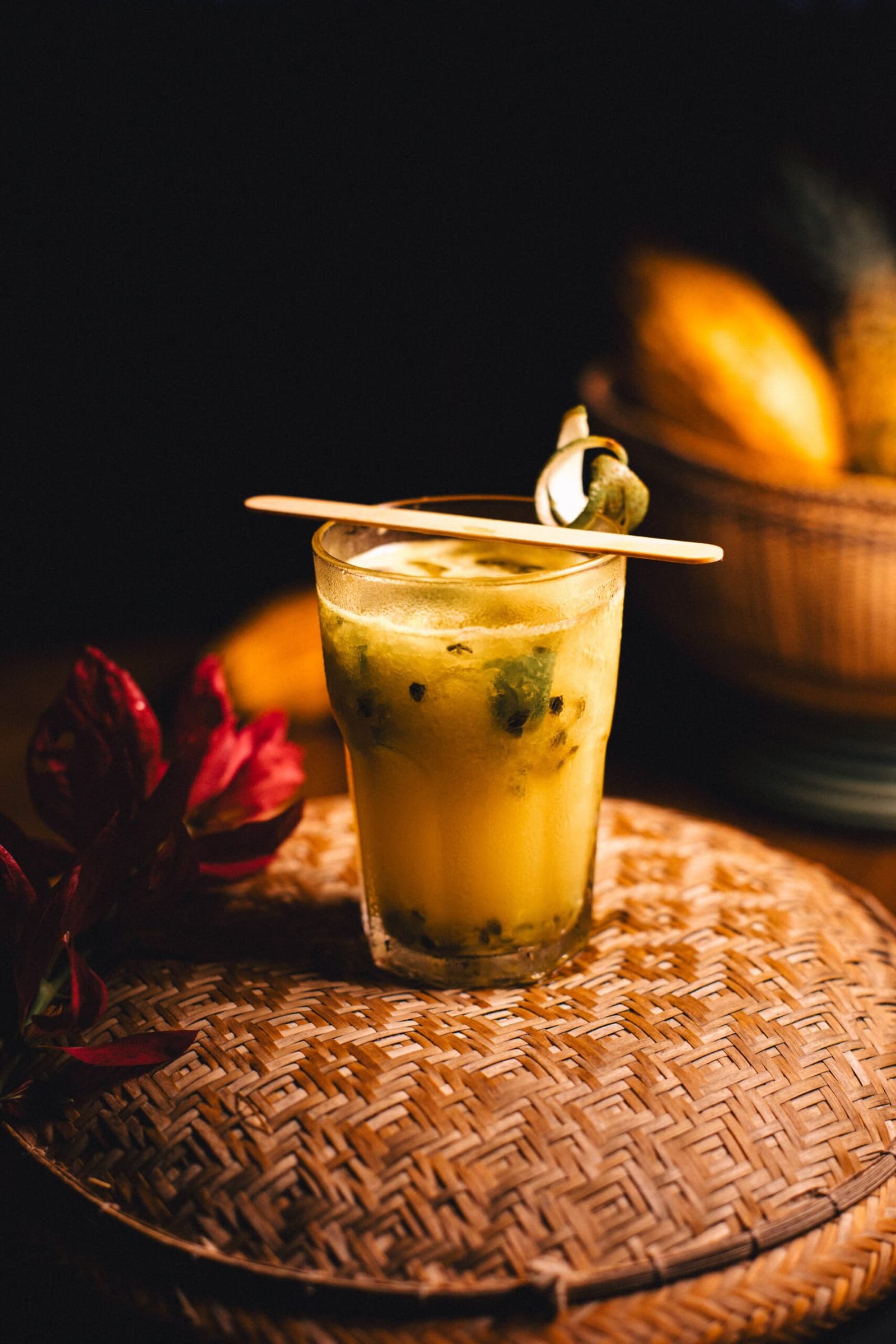 Ein Schluck, und der Regenwald fühlt sich schon vertrauter an. Ein goldenes Maracuja-Cocktail mit sichtbaren Kernen in einem Glas auf einem geflochtenen Rattantablett in der Anavilhanas Jungle Lodge Brazil, garniert mit einem Holzrührstab und einem eingerollten Tropenblatt, im Hintergrund ein Korb mit gelben Früchten