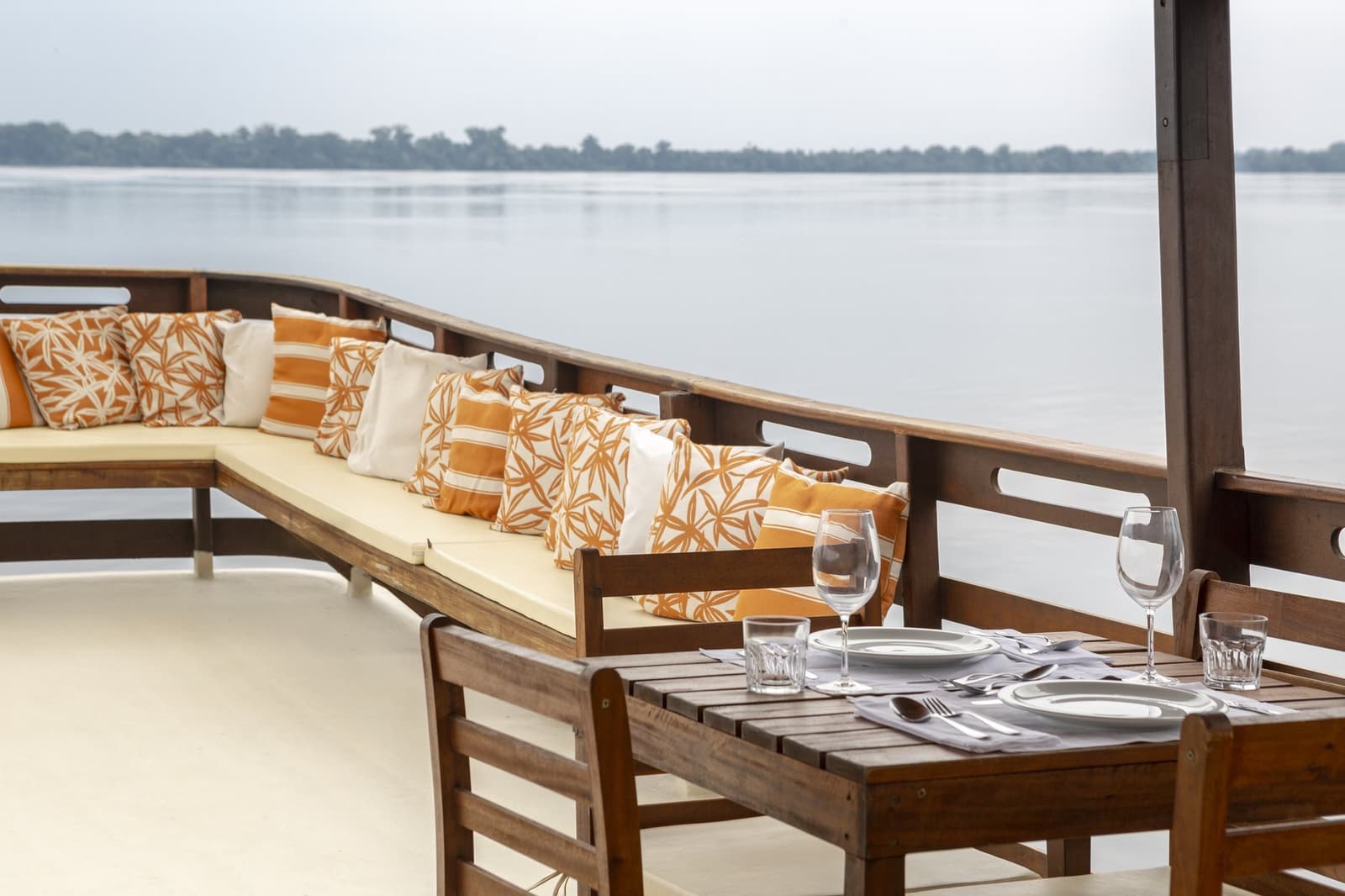 Abendessen auf dem Fluss, während der Regenwald am Horizont wartet. Gedeckter Esstisch mit Weingläsern und Besteck auf dem offenen Deck eines Bootes im Anavilhanas Jungle Lodge Brazil, mit einem gepolsterten Loungesofa und einem breiten Fluss vor bewaldeter Kulisse