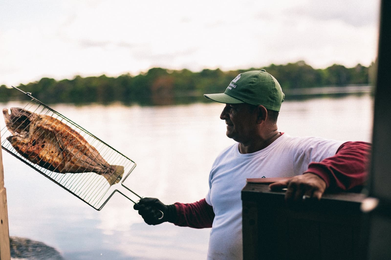 Frisch aus dem Fluss, direkt auf den Rost. Kochen, das keine großen Worte braucht. Ein lächelnder Mann mit grüner Kappe hält einen ganzen gegrillten Fisch in einem Drahtkorb über dem Fluss, mit dichtem Regenwald im Hintergrund