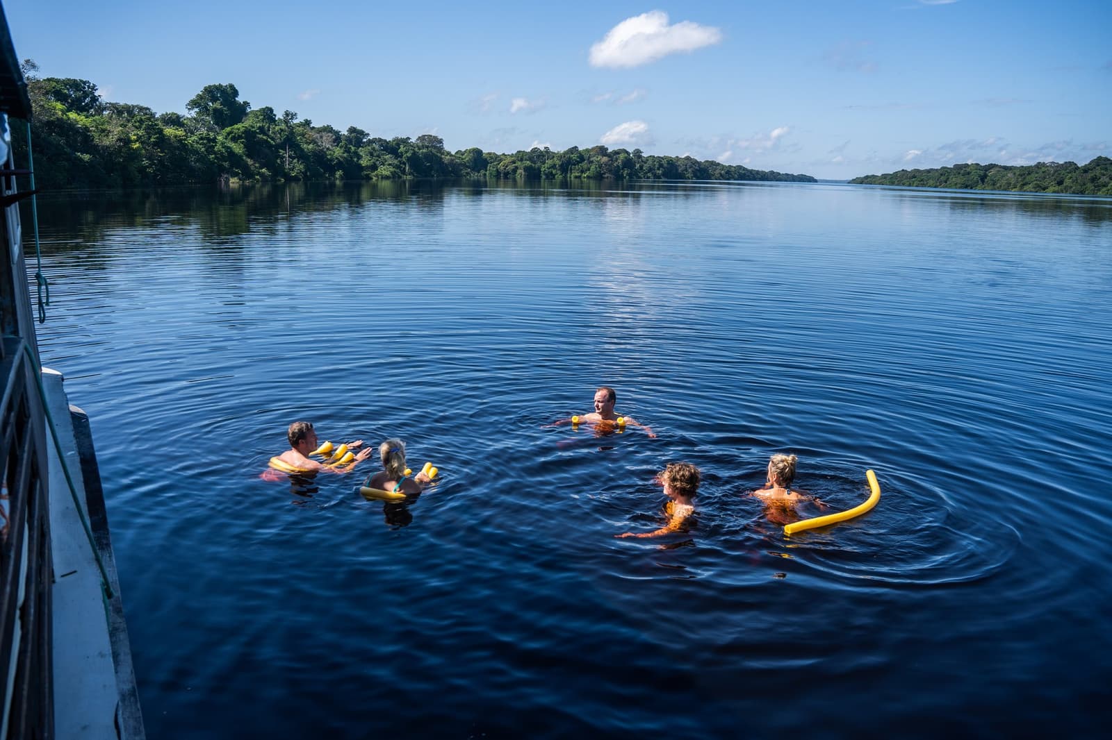 Schwarzes Wasser, blauer Himmel, und plötzlich schwimmst du mitten im Amazonas. Fünf Gäste schwimmen im dunklen Wasser des Rio Negro neben einem traditionellen Boot der Anavilhanas Jungle Lodge Brazil, mit gelben Schwimmnudeln und dichtem Regenwald an beiden Ufern unter blauem Himmel
