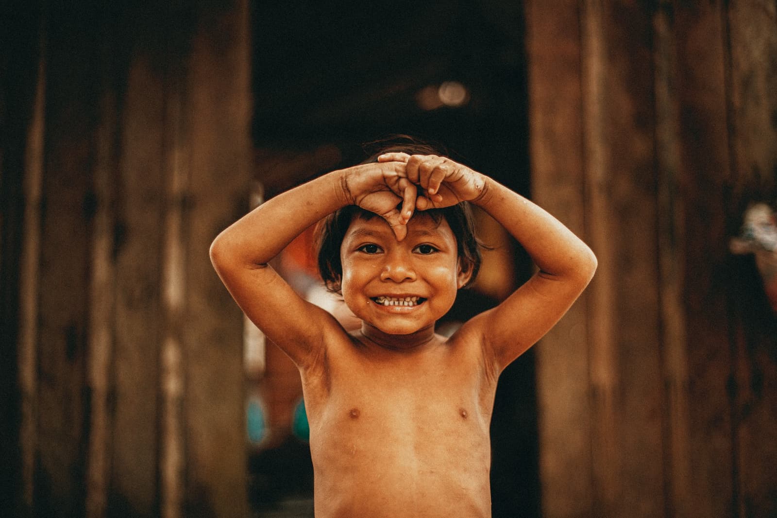 Manche Momente passieren einfach, ohne Vorwarnung. Ein lachendes Kind in einer Ufergemeinde nahe der Anavilhanas Jungle Lodge Brasilien, mit verspielt erhobenen Händen vor einer Holzwand