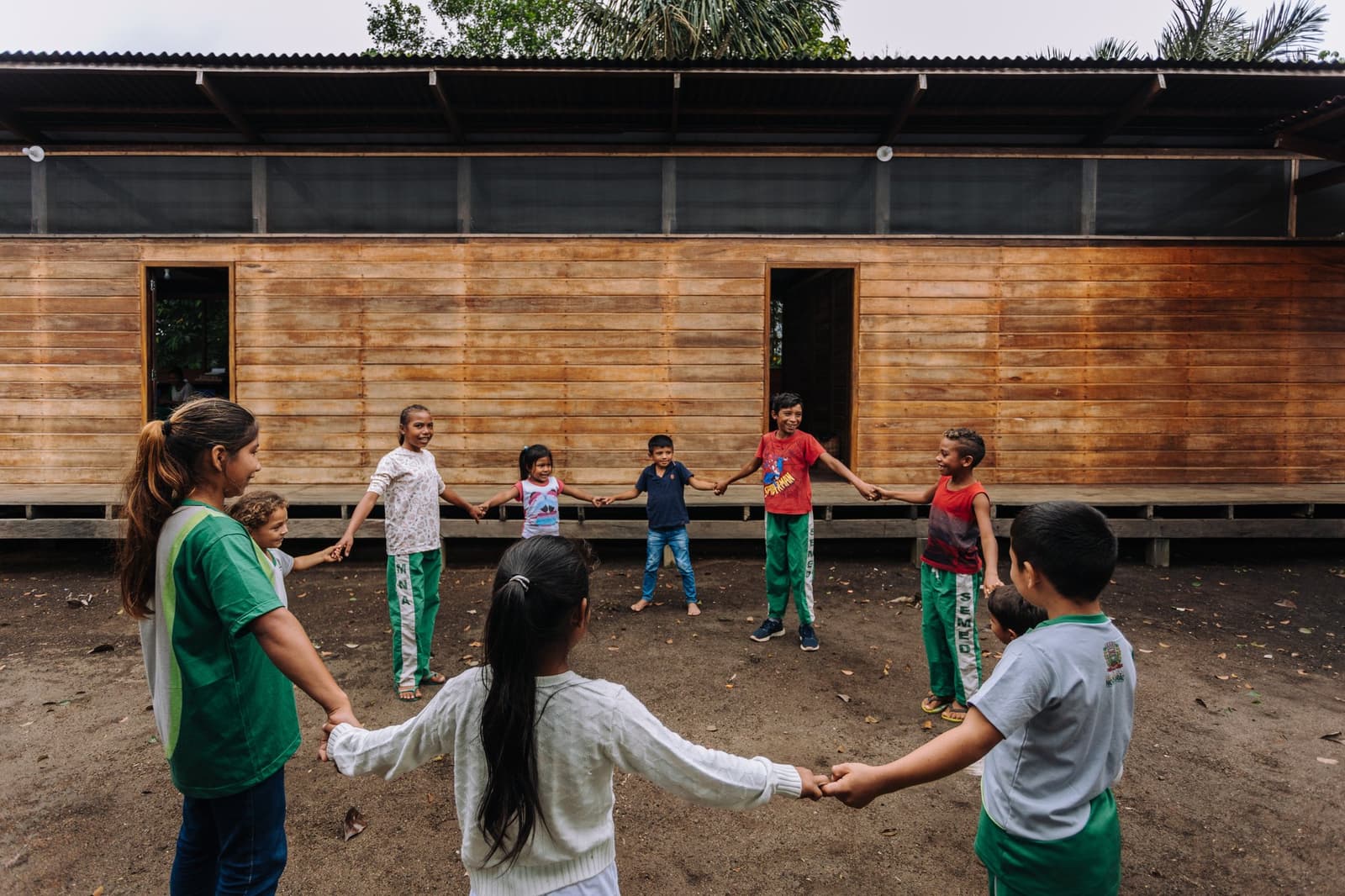 Ein Kreis, viele Lachen, und der Wald hört einfach zu. Kinder halten sich an den Händen und spielen im Kreis vor einem Holzgebäude bei einem Gemeinschaftsbesuch in der Anavilhanas Jungle Lodge Brazil