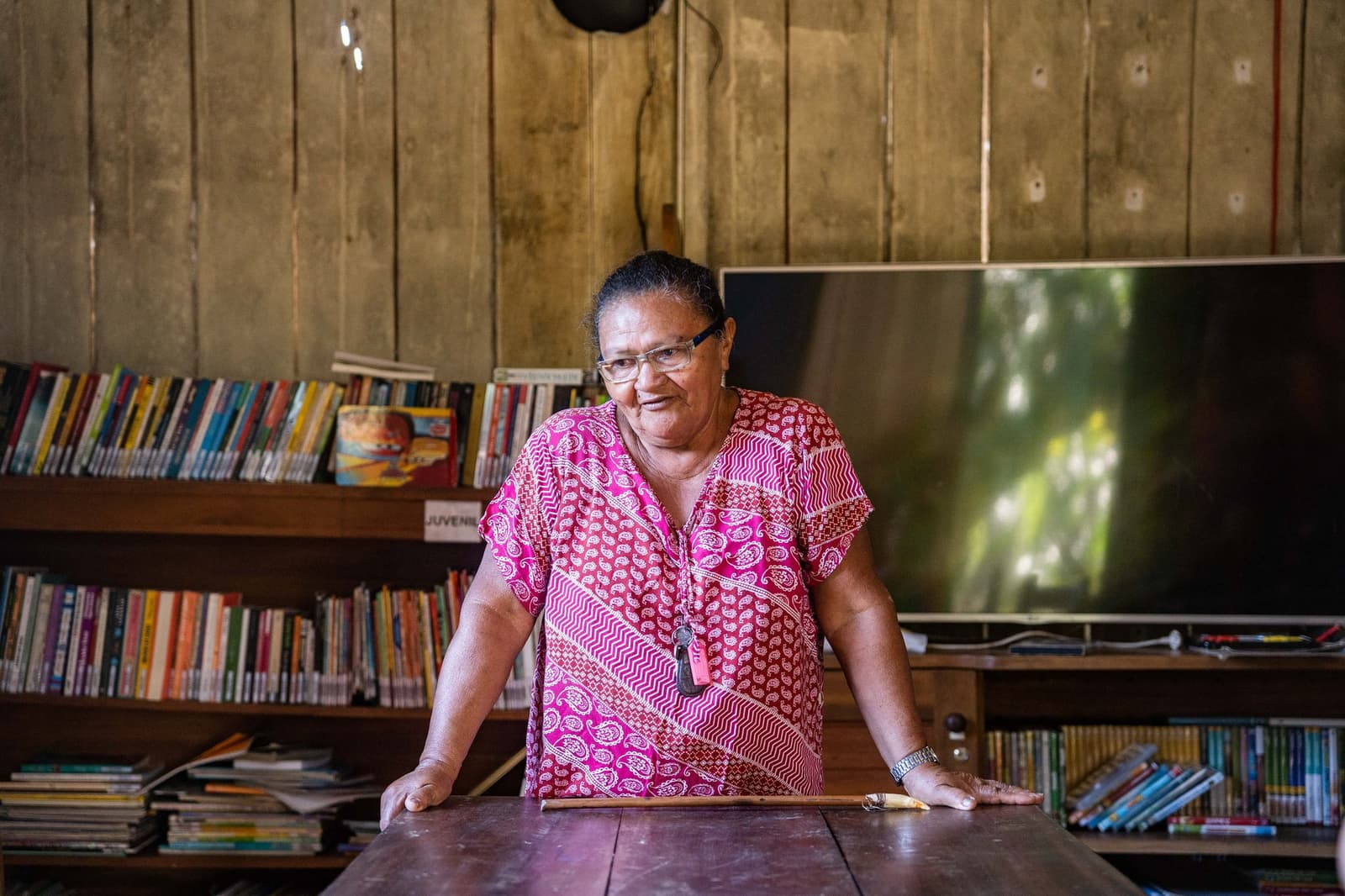 Sie kennt jedes Buch im Regal, und wahrscheinlich auch jeden, der es ausgeliehen hat. Eine einheimische Frau in einem gemusterten rosa Kleid steht an einem Holztisch in einer Gemeindebibliothek in der Nähe der Anavilhanas Jungle Lodge Brazil, hinter ihr bunte Bücherregale an Holzwänden