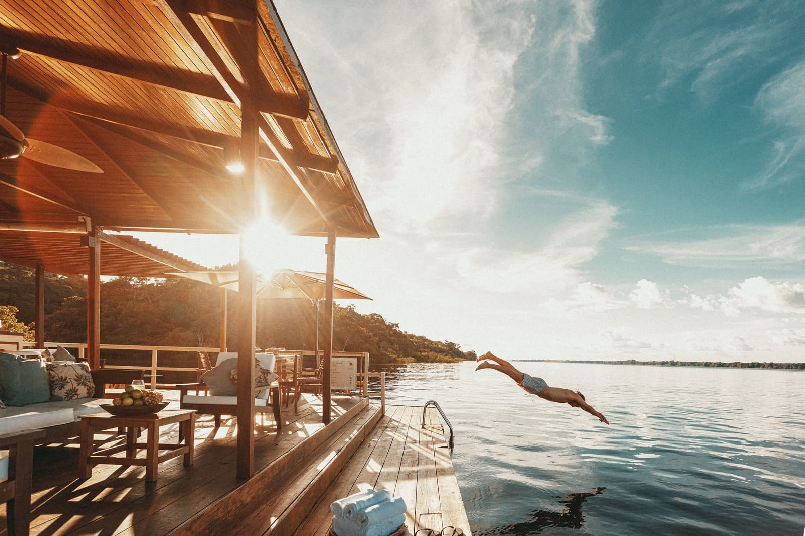 Kein langer Anlauf nötig, nur ein kurzer Schritt vom Steg. Ein Gast springt vom Holzdeck der Anavilhanas Jungle Lodge Brazil in den stillen Rio Negro im goldenen Abendlicht, während ein möblierter Loungebereich und üppiger Regenwald im Hintergrund zu sehen sind