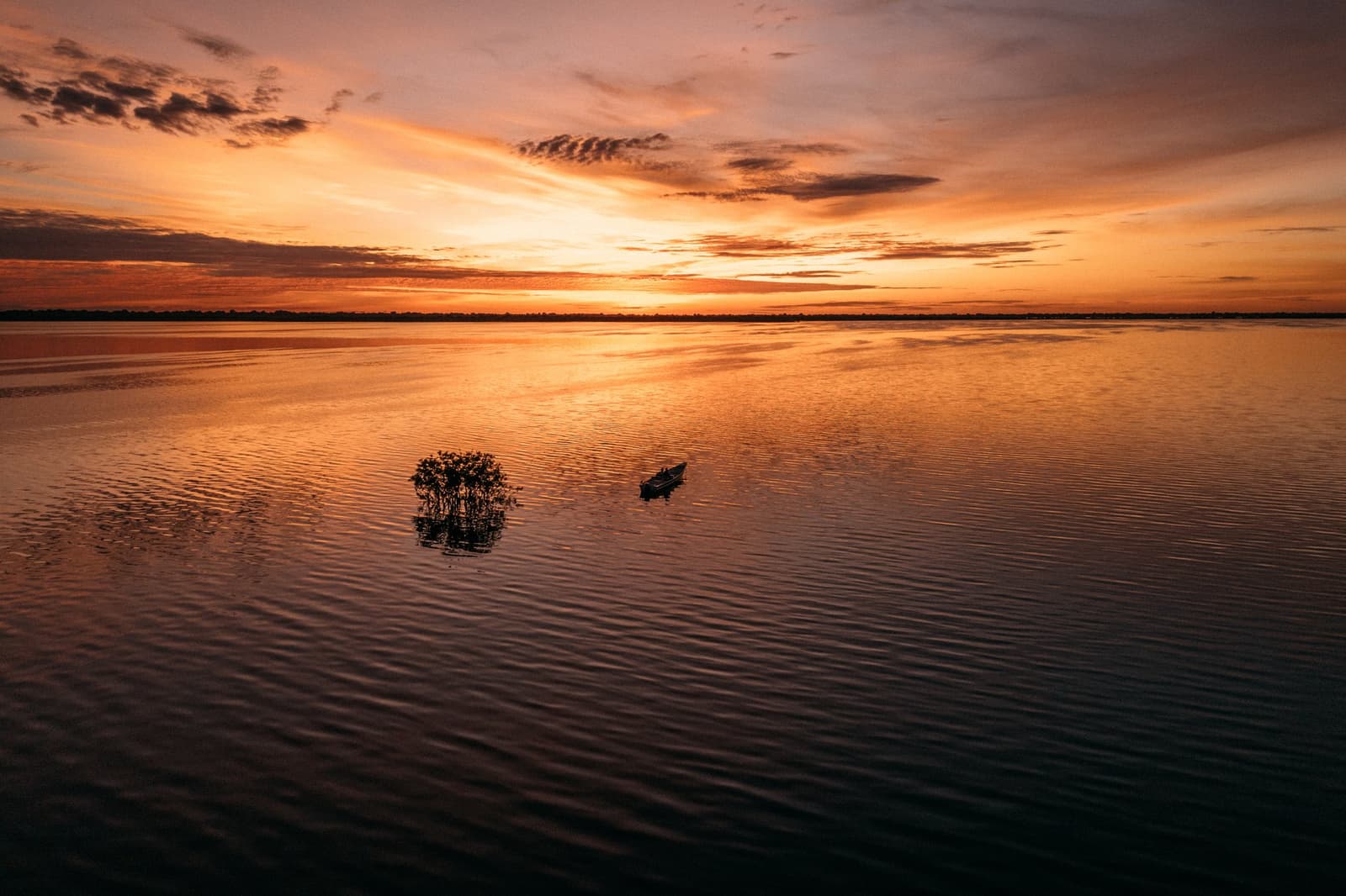 Ein Kanu, ein versunkener Baum und der halbe Himmel als Spiegel, mehr braucht es nicht. Ein kleines Motorboot gleitet bei Sonnenaufgang über den spiegelglatten Rio Negro nahe der Anavilhanas Jungle Lodge Brazil, ein einzelner überfluteter Strauch zeichnet sich gegen den orange-roten Himmel ab