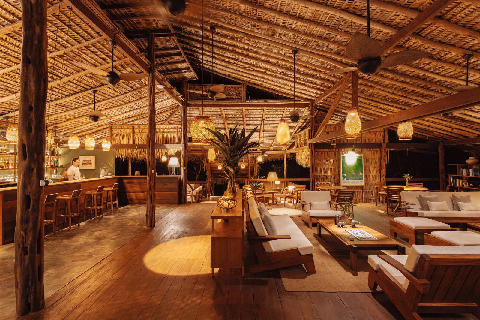 Hier vergeht der Abend schneller als ein Sonnenuntergang am Rio Negro. Warmbeleuchtete Open-Air-Lounge des Anavilhanas Jungle Lodge Brazil mit Palmstrohdach, geflochtenen Hängeleuchten, Hartholzböden, Leinensofas und einer Holzbar im Hintergrund
