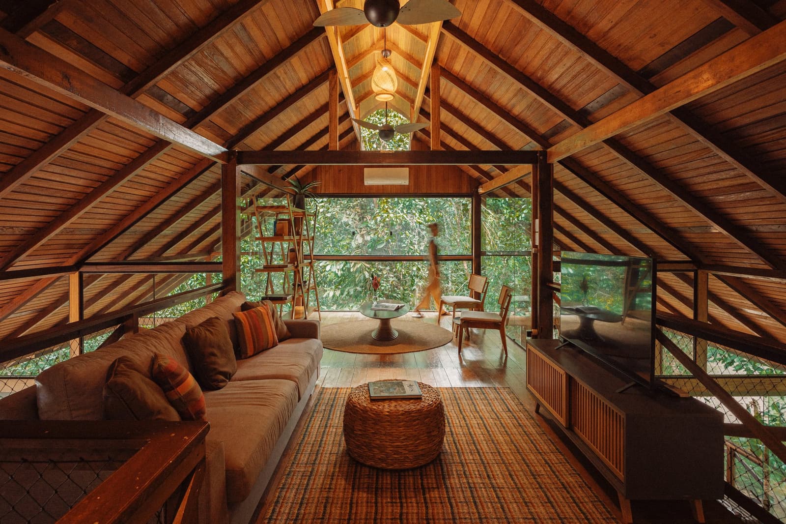 Ein Wohnzimmer, das jeden Stadtblick vergessen lässt. Gewölbte Holzdecke und offener Wohnbereich der Anavilhanas Jungle Lodge Brasilien mit Sofa, gewebtem Teppich, Rattanhocker und Panoramablick auf den Regenwald