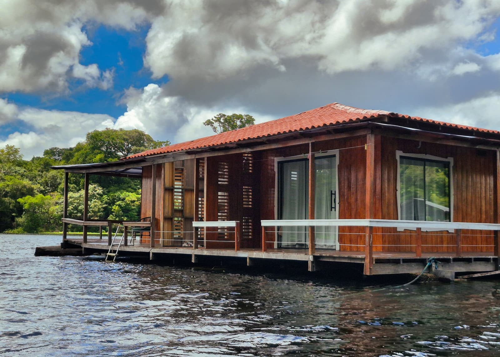 Holzwände, ein eigenes Deck und rundum Dschungelstille — schlafen mitten im Regenwald. Ein hölzernes Schwimmlodge auf dunklem Flusswasser mit umlaufender Terrasse, Jalousieläden und Glasschiebetüren, eingebettet in üppigen Regenwald — Teil einer Brasilien-Amazonas-Dschungeltour