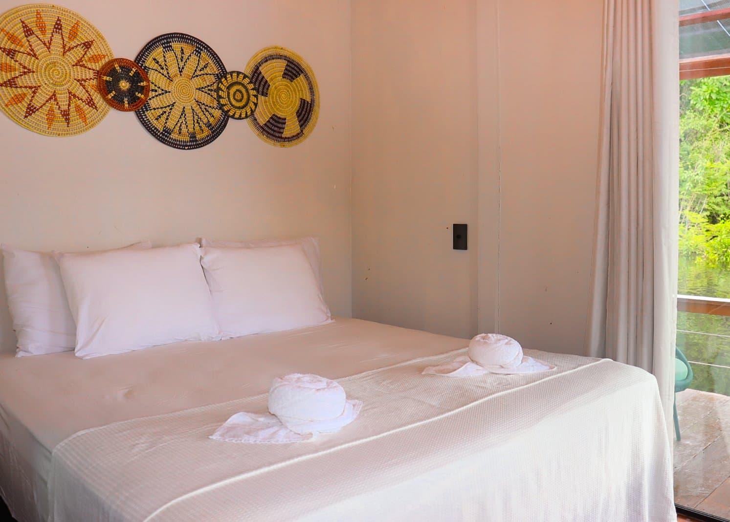 Handgemachte Wanddeko, frische Bettwäsche – und der Regenwald direkt vor dem Fenster. Ein Doppelbett mit weißer Bettwäsche und gefalteten Handtüchern unter gewebter Korbwandkunst in einem gemütlichen Lodgezimmer, mit Blick auf den grünen Dschungel