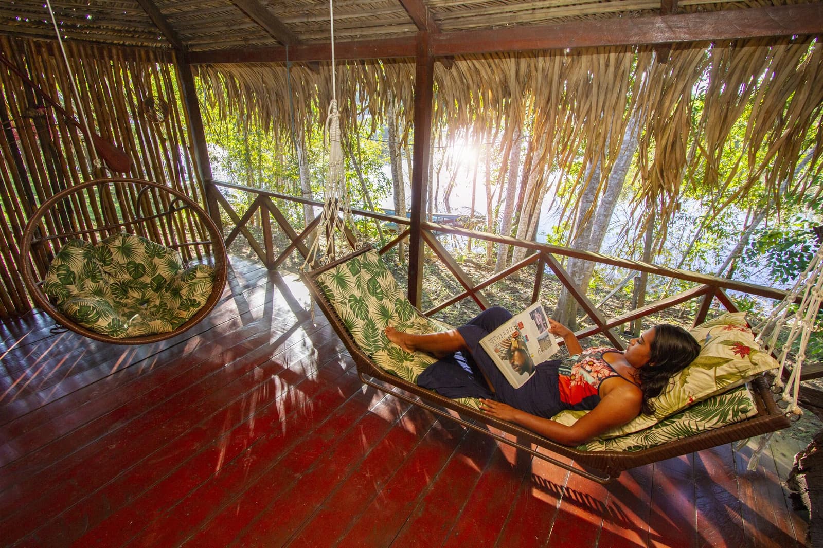 Ein Schaukelbett, goldenes Licht und der Dschungel zum Greifen nah. Photo: David Nascimento @ Eine Gästin liegt in einem Rattan-Schaukelbett auf einer Veranda mit Strohdach und liest ein Magazin, während Sonnenlicht durch den tropischen Wald im Hintergrund fällt