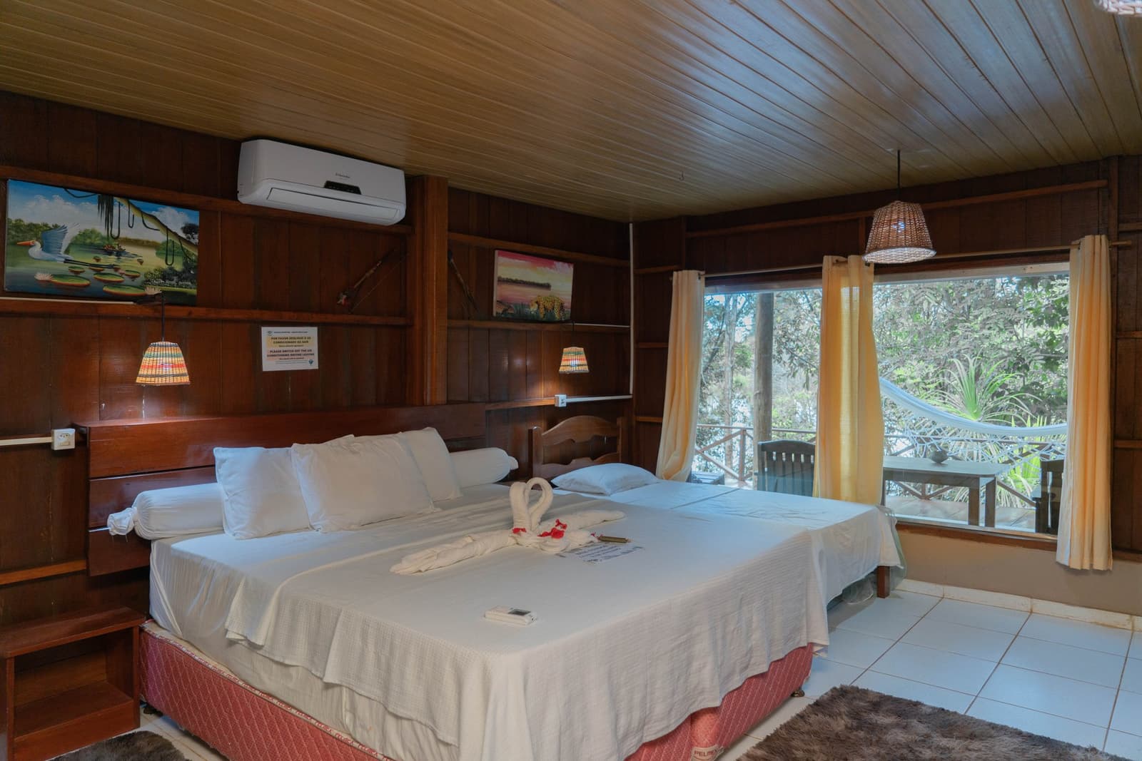 Handtuch-Schwäne auf weißen Laken — der Dschungel wartet direkt hinter dem Fenster. Photo: Yuri Panichi Komfortables Doppelzimmer in einer Dschungellodge mit dunklen Holzwänden, Wildtier-Kunstwerken, geflochtenen Lampenschirmen und einem privaten Balkon mit Hängematte, von dem aus man in den Regenwald schaut